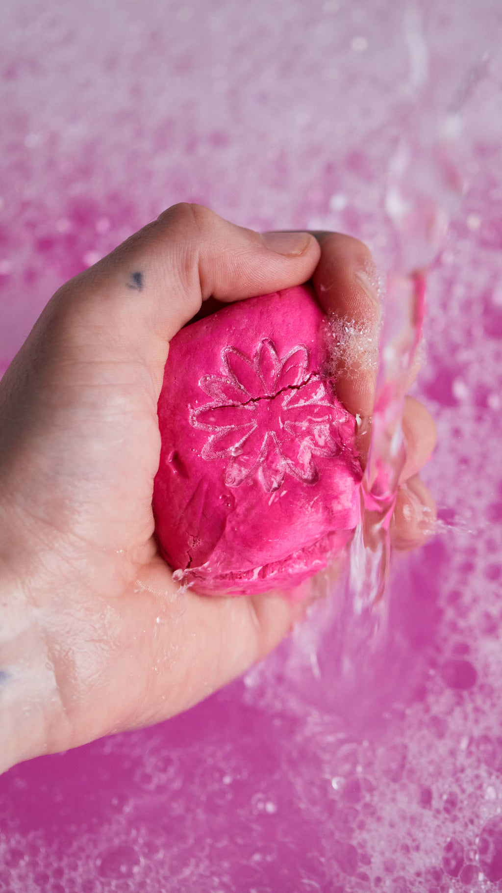 Image shows the model gently crumbling the pink bubble bar under running water in front of bubbly, pink bath water.
