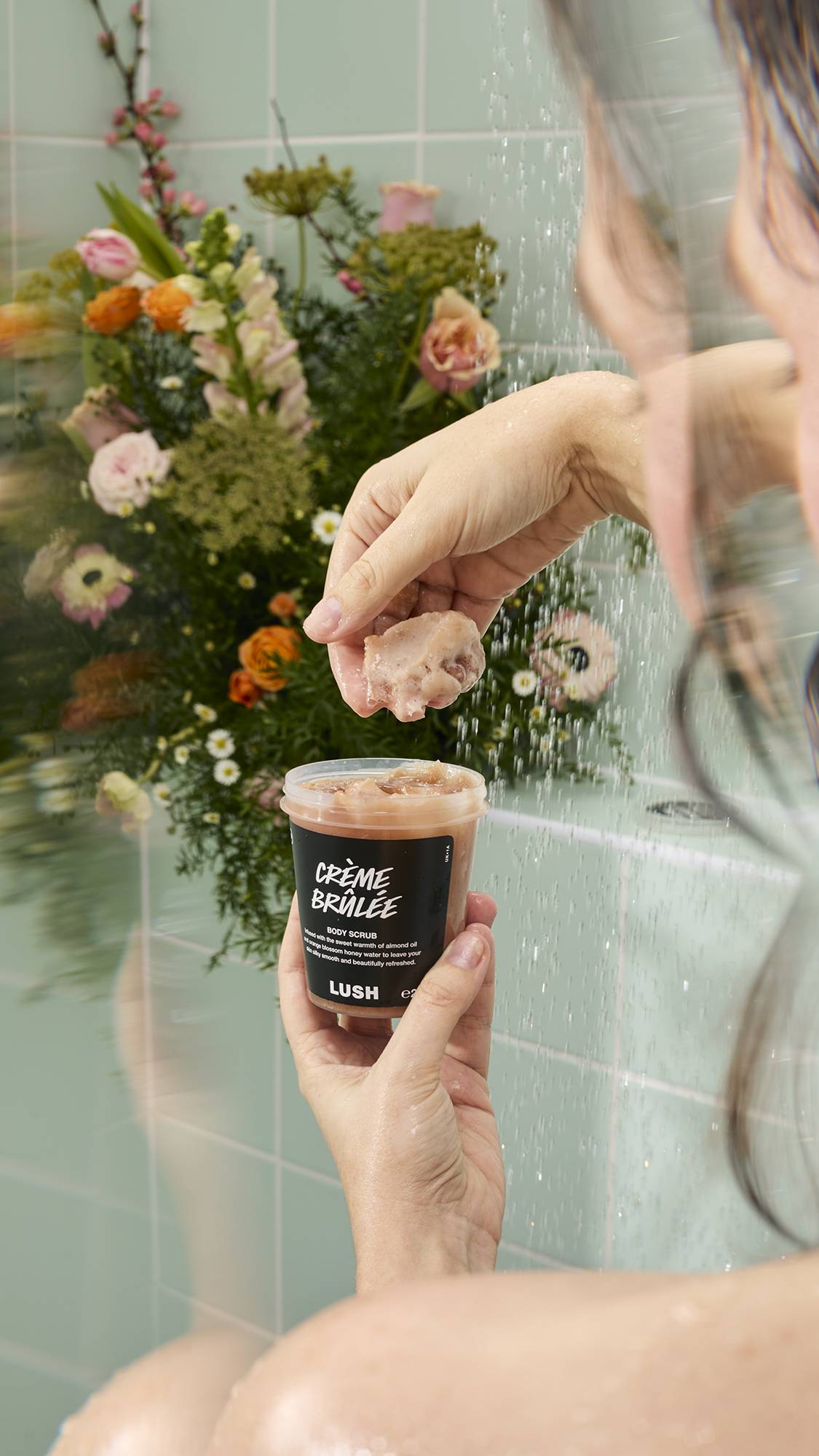 A person in a shower holding a pot of Lush Crème Brûlée body scrub, scooping out a handful of the thick, caramel-coloured product. A large, colourful bouquet of flowers is visible in the background against light green bathroom tiles.