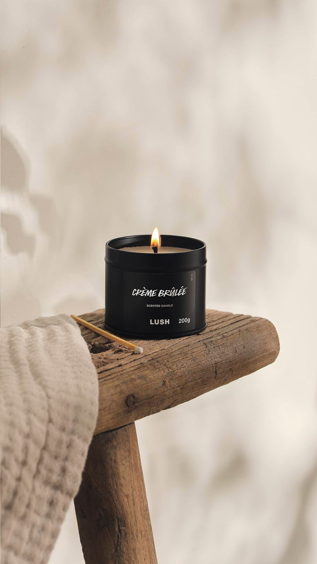 The image shows a blurred, cream background. In the foreground, the Creme Brûlée black tin candle sits with a lit flame on a rustic wooden stool with an unused match beside it and a cream cloth.