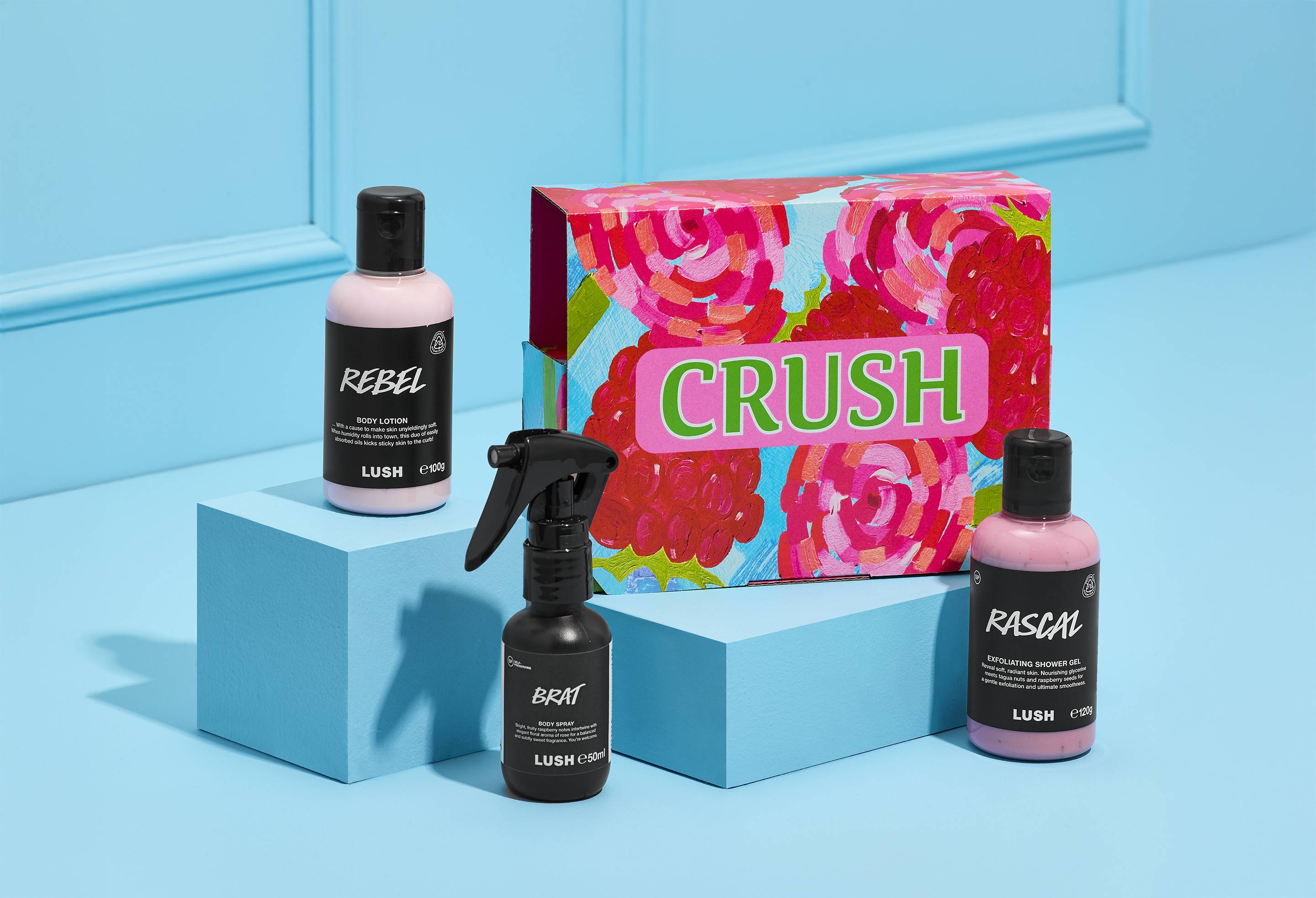 A LUSH Crush gift set displayed on bright blue risers against a matching blue backdrop. The set includes a pink rose-patterned "CRUSH" box, "Rebel" body lotion, "Rascal" shower gel, and "Brat" body spray.
