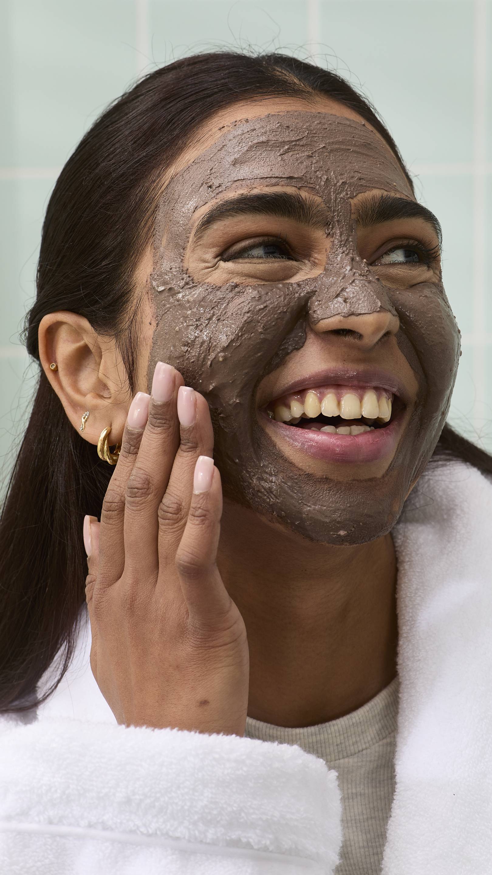 A model is smiling broadly while applying a thick, dark brown "Cupcake" face mask to their forehead and cheeks. They are wearing a white dressing gown and gold hoop earrings, set against a background of pale green bathroom tiles.
