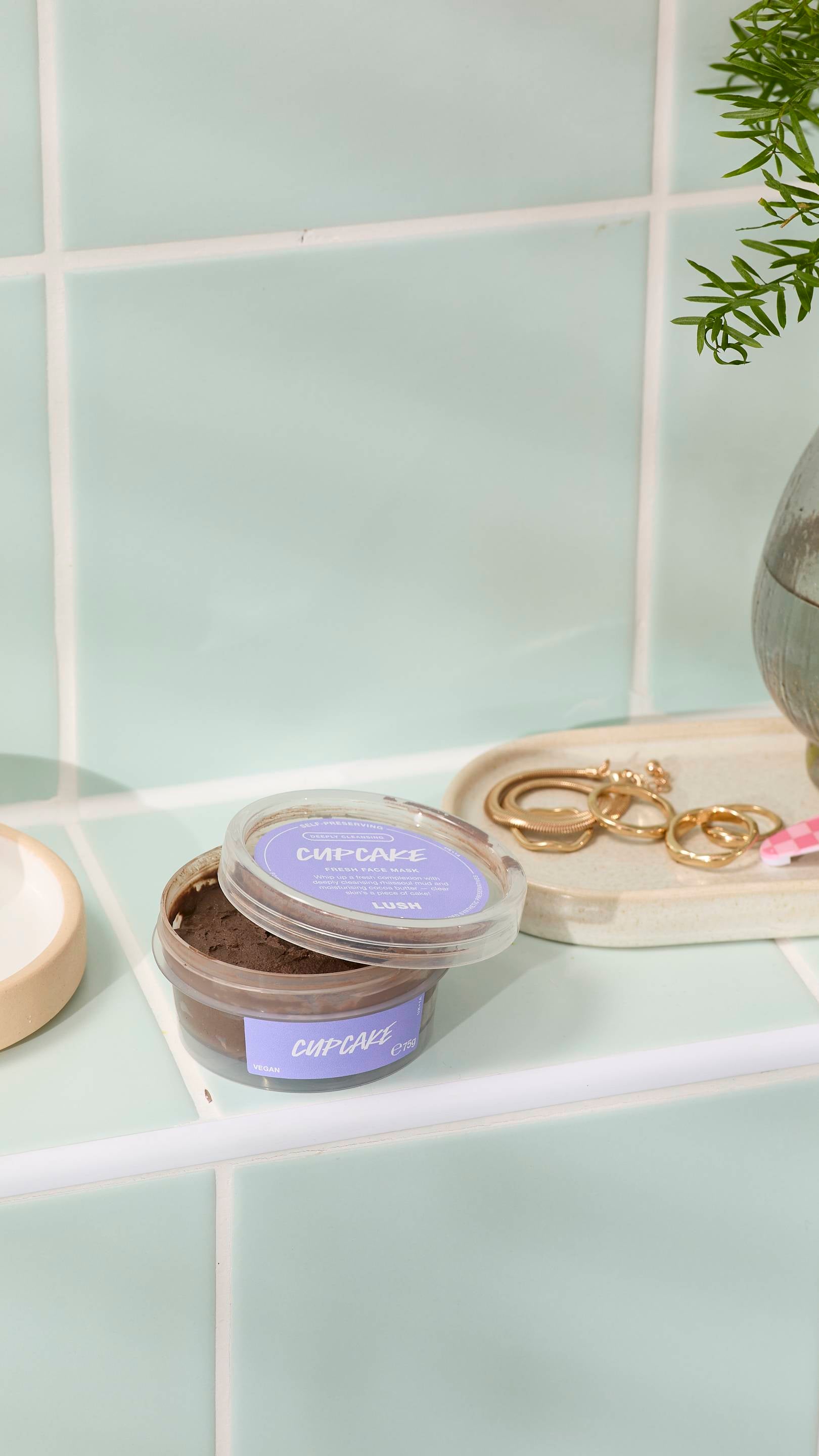 An open pot of dark brown "Cupcake" fresh face mask sits on pale green bathroom tiles. In the background, a small tray holds gold rings and hoop earrings, next to a glass vase with a green plant.