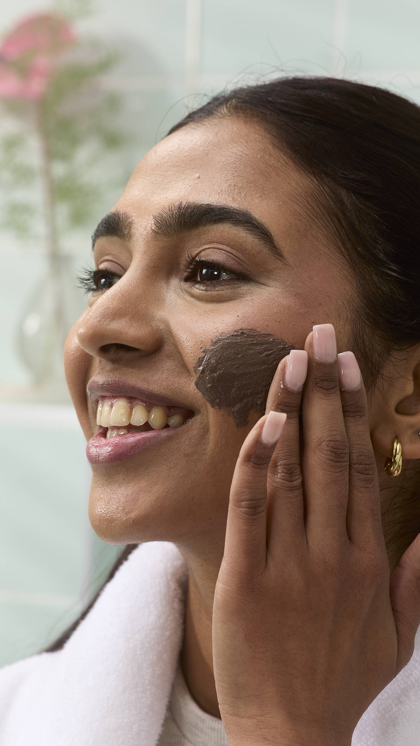 A close-up of a model smiling while applying a dark brown "Cupcake" face mask to their cheek with their hand. They are wearing a white dressing gown, set against a blurred bathroom background.