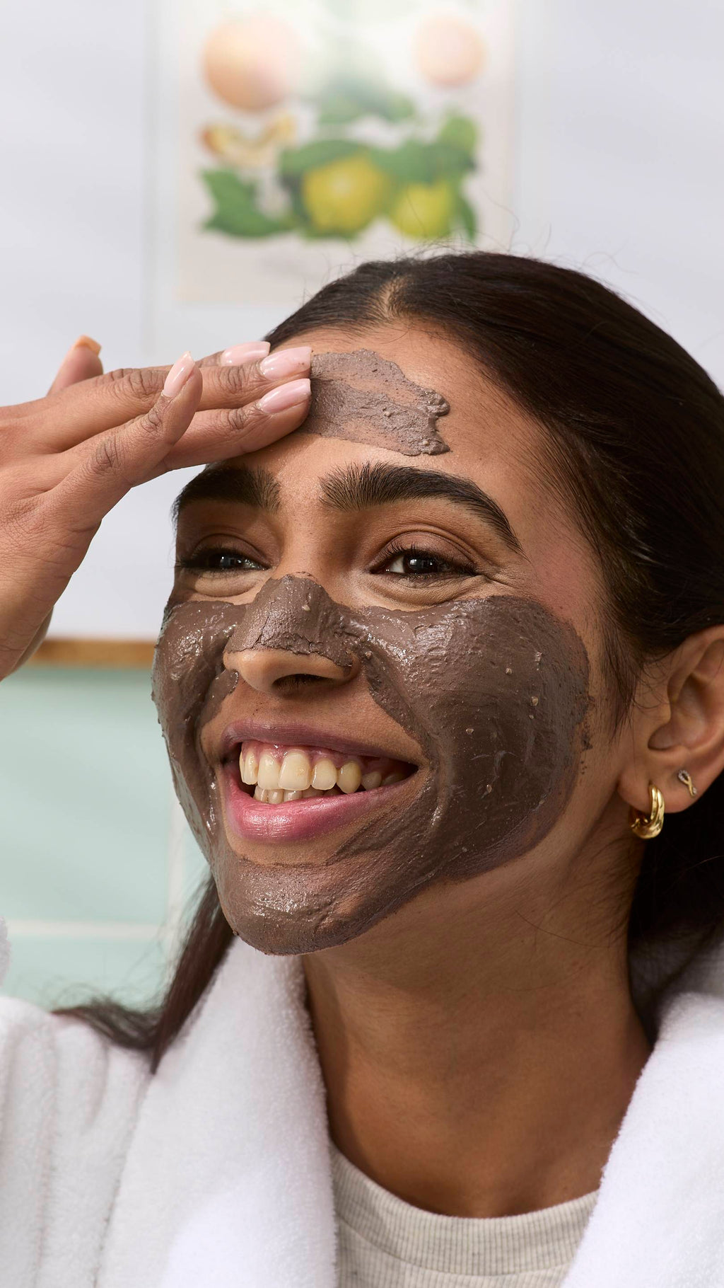 A close-up of a model smiling while applying a dark brown "Cupcake" face mask to their forehead with their hand. They are wearing a white dressing gown, set against a blurred bathroom background.