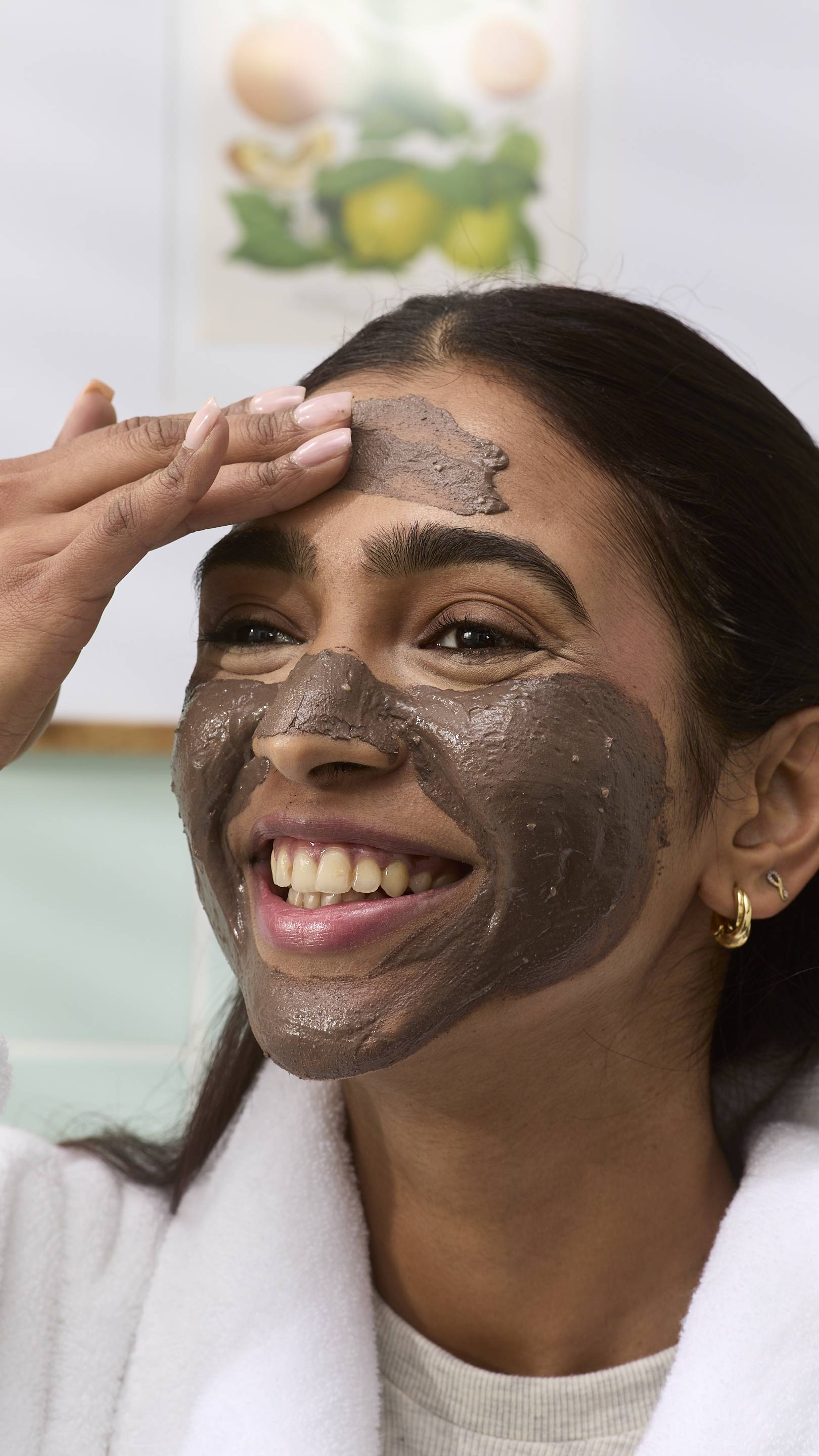 A close-up of a model smiling while applying a dark brown "Cupcake" face mask to their forehead with their hand. They are wearing a white dressing gown, set against a blurred bathroom background.