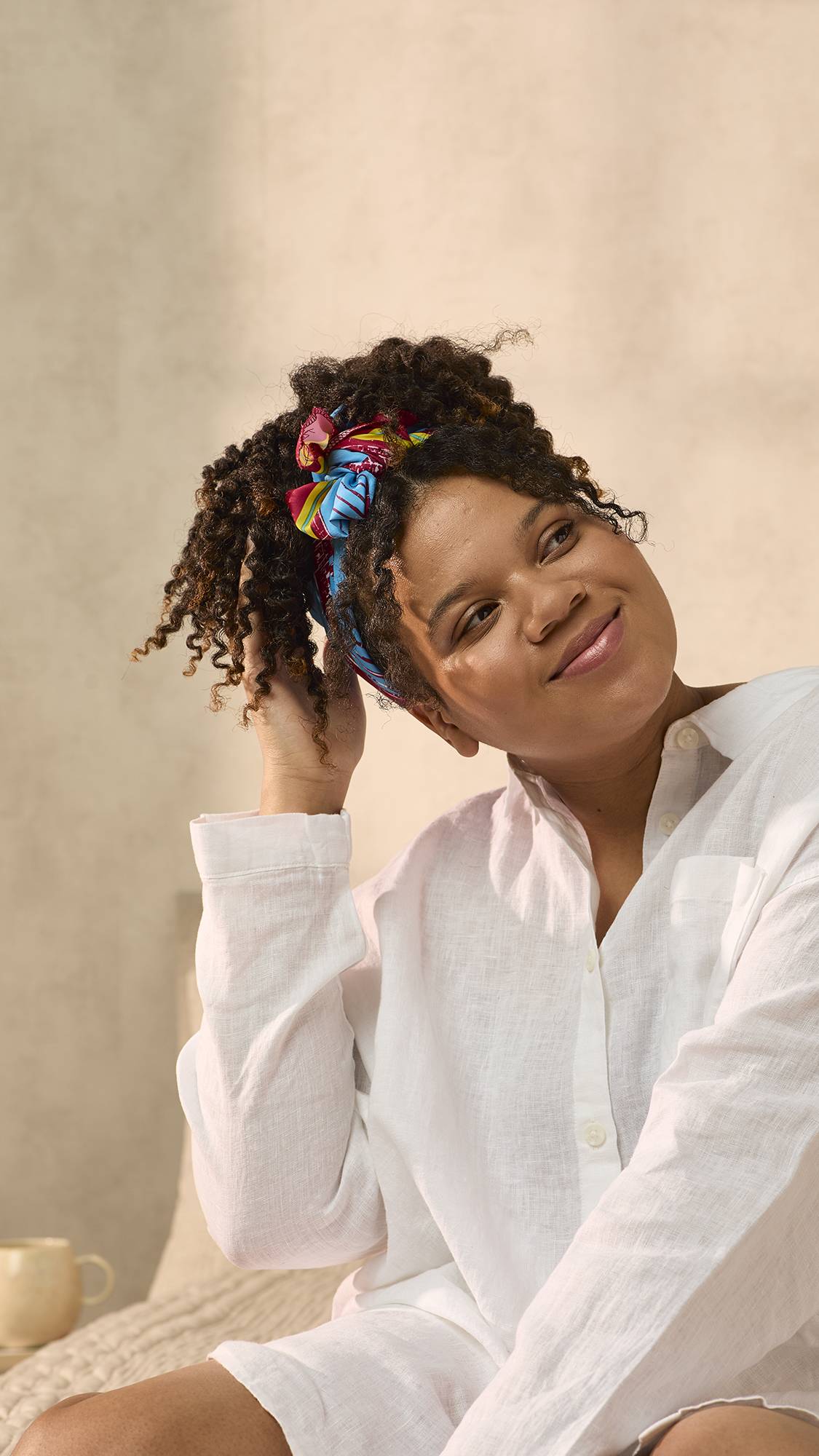 The model is sat on the bed in an oversized white shirt. They have fashioned the Curly Coily gift knot wrap into a trendy head bandana for their thick, curly hair.
