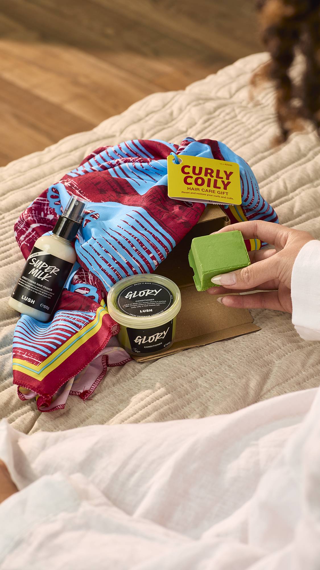 The image shows the Curly Coily gift, having been unpacked, with the three included haircare products on the bed.