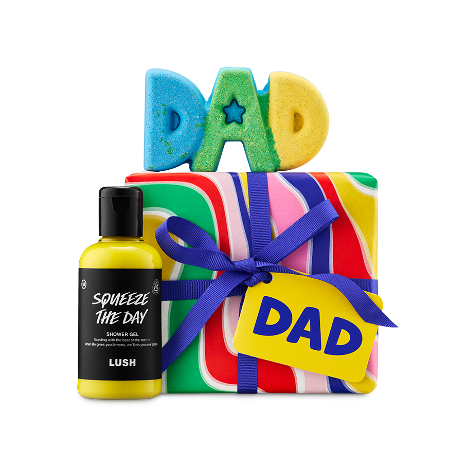 DAD gift. A vibrant Lush gift set featuring a colourful swirl-patterned gift box with a blue ribbon and "DAD" tag, and a blue, green, and yellow "DAD" shaped bath bomb on top and "Squeeze The Day" shower gel to the side.