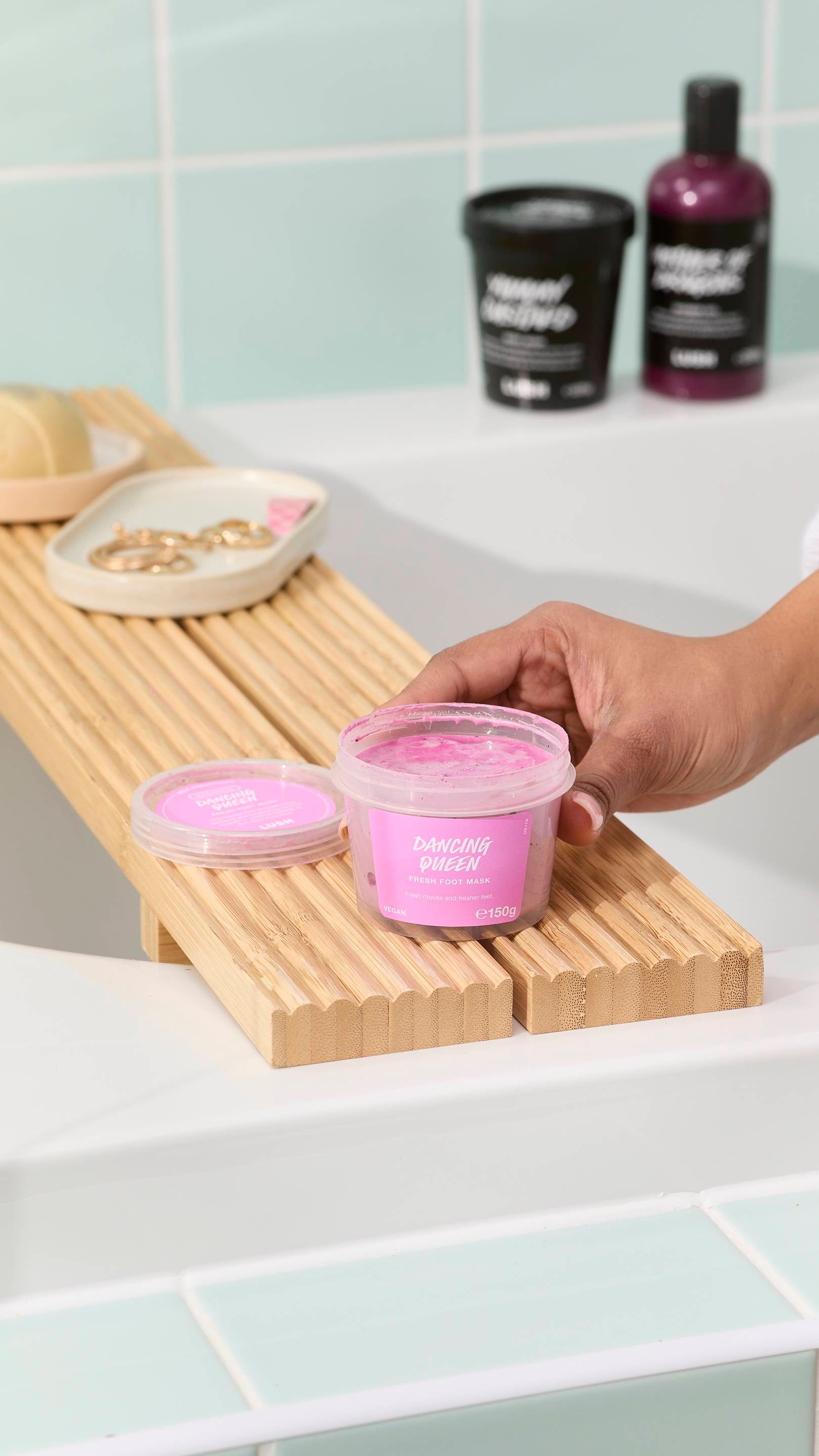 A hand reaches for an open tub of "Dancing Queen" pink foot mask sitting on a wooden bath tray. The background features a white bathtub, light green wall tiles, and other LUSH self-care products.