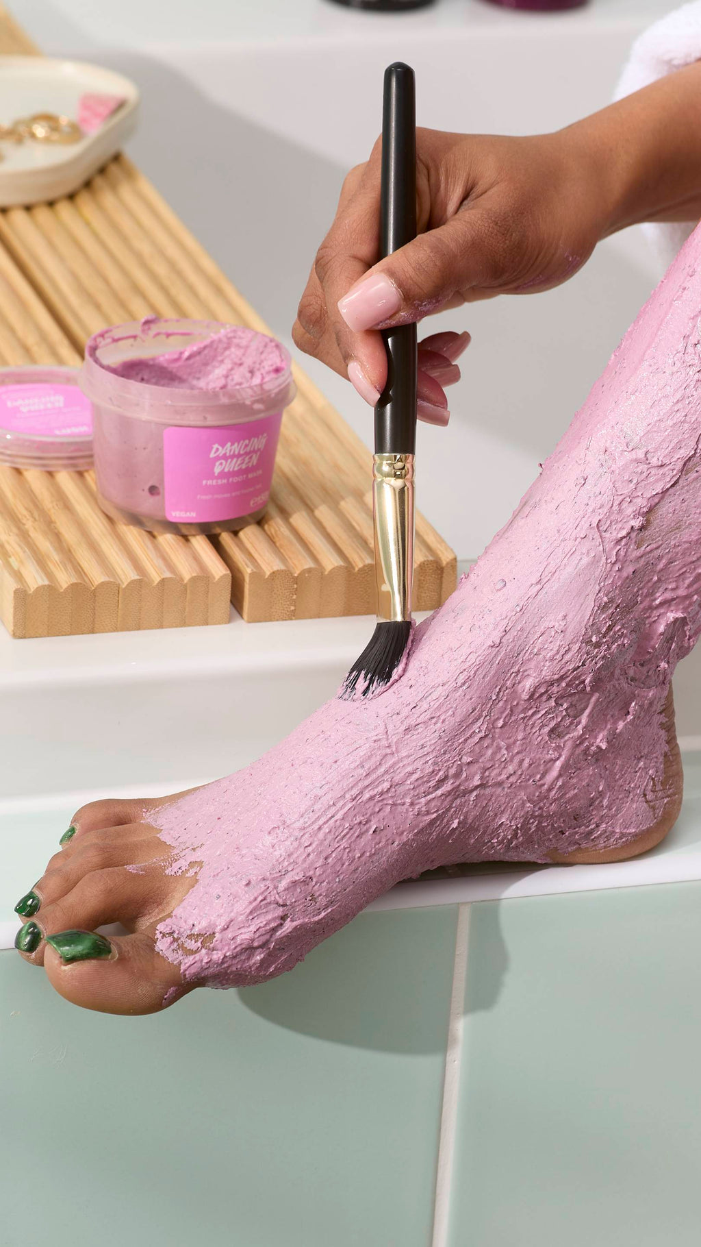 A close-up of a person using a black-handled brush with gold detailing to apply a thick pink foot mask to their foot. The "Dancing Queen" foot mask tub sits on a wooden tray in the background against light green tiles.