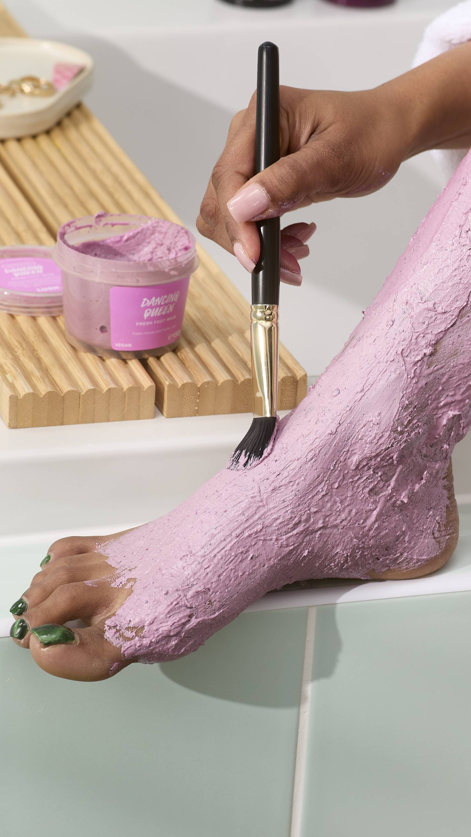 A close-up of a person using a black-handled brush with gold detailing to apply a thick pink foot mask to their foot. The "Dancing Queen" foot mask tub sits on a wooden tray in the background against light green tiles.
