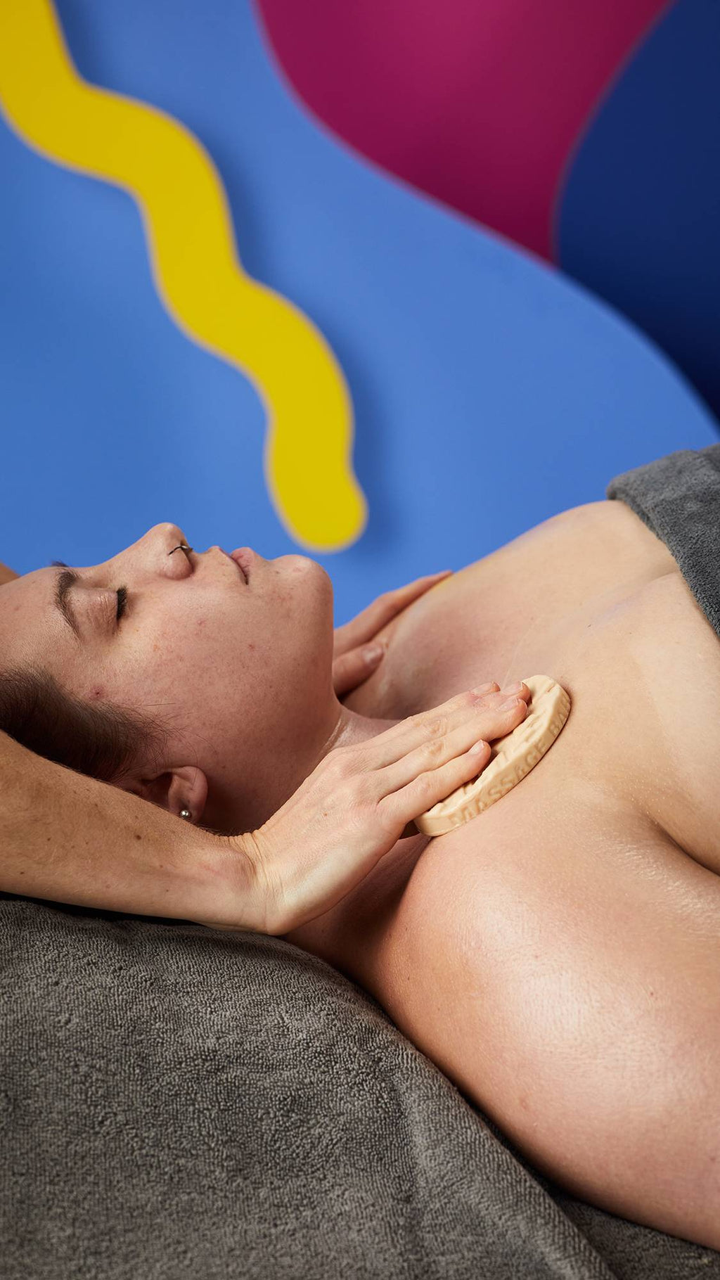 A person lies with eyes closed on a grey towel as hands glide an oval, beige Lush massage bar across their collarbone. Set against a backdrop of bright blue, pink, and yellow abstract shapes.