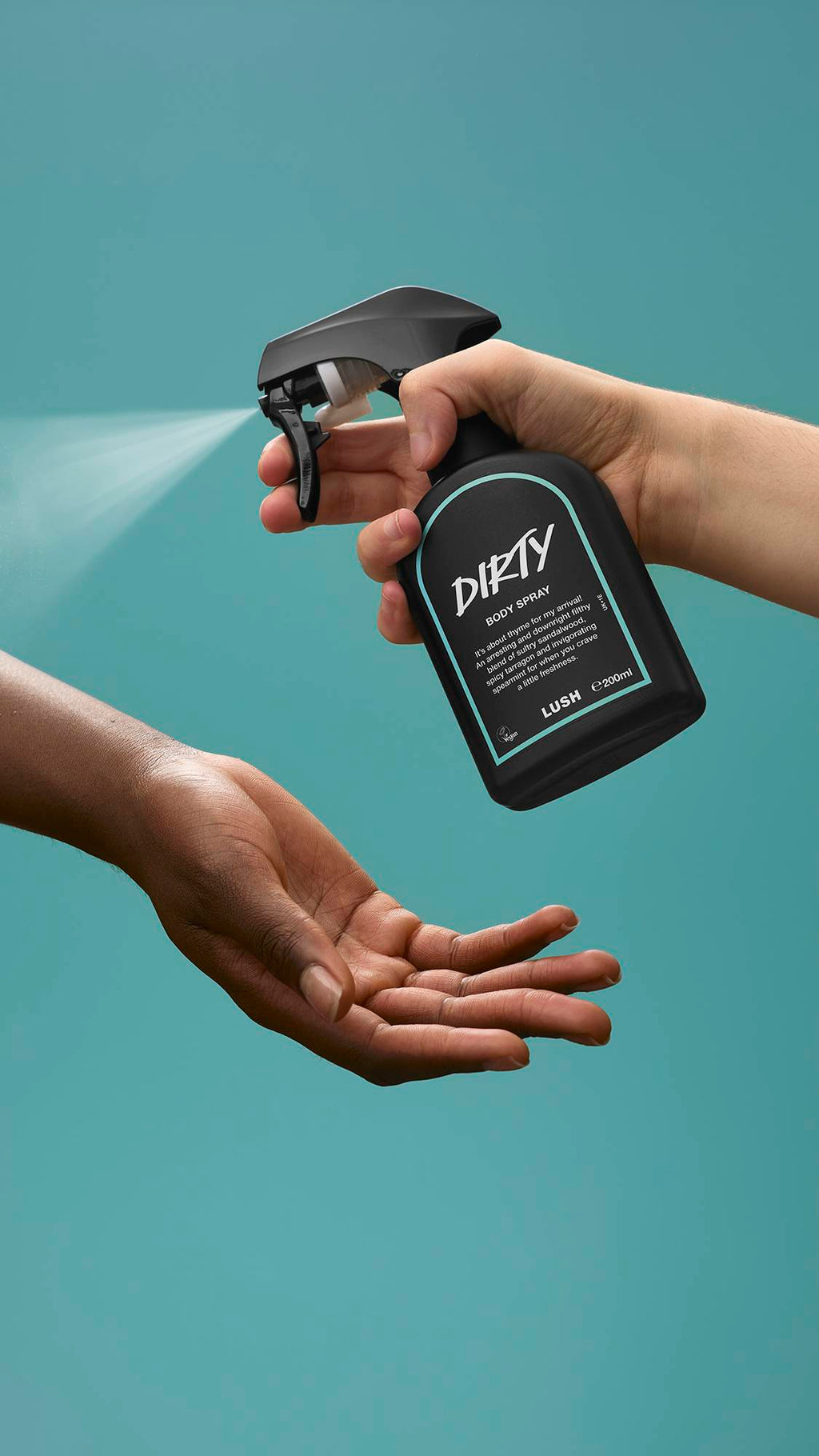 One hand holds a black bottle of Lush 'Dirty' body spray and spritzes a fine mist over another person's open, waiting hand. Set against a solid teal-blue background.