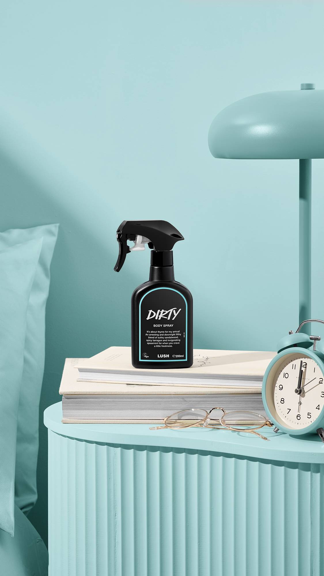 A black bottle of Lush 'Dirty' body spray sits on a stack of books on a ribbed, pastel blue bedside table. Next to it are gold-rimmed glasses, a matching blue alarm clock, and a lamp, creating a colour-coordinated bedroom scene.