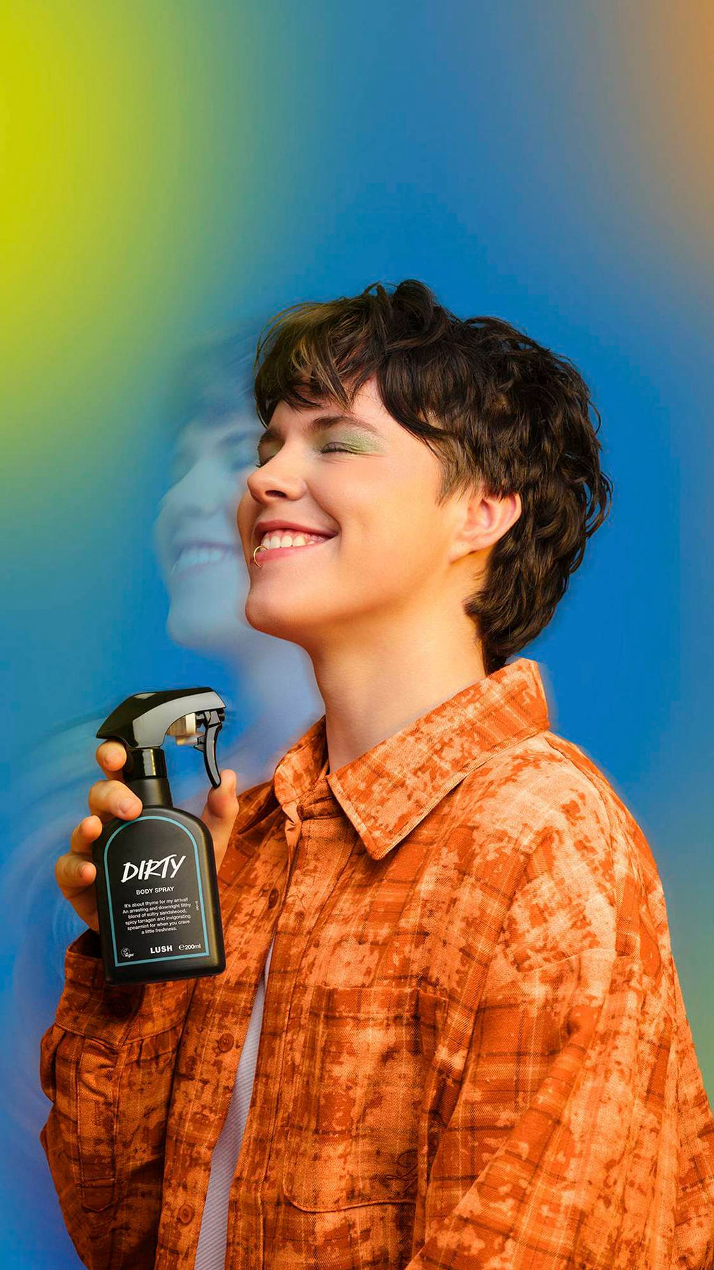 A smiling person with short wavy hair holds a black Lush 'Dirty' body spray. They wear an orange patterned shirt. A motion-blurred double exposure effect of their face floats behind them against a blue and yellow gradient background.