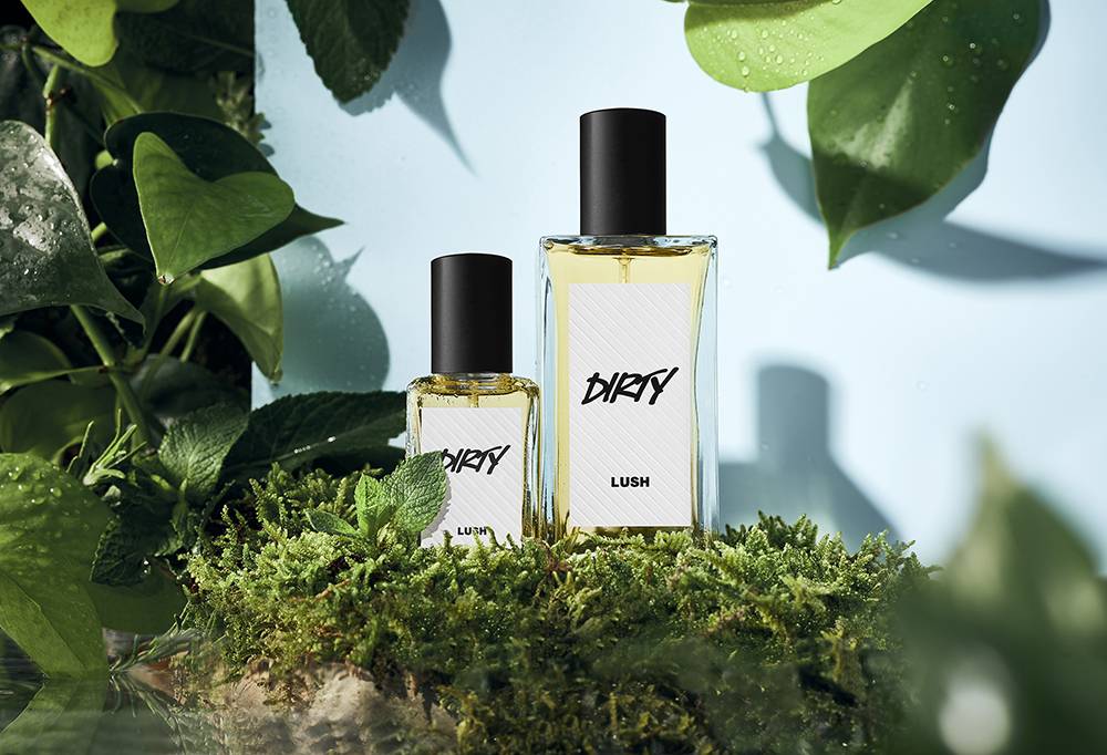 Dirty perfume is placed on green grass. Green leaves with water droplets are draped in the corners on a blue backdrop.