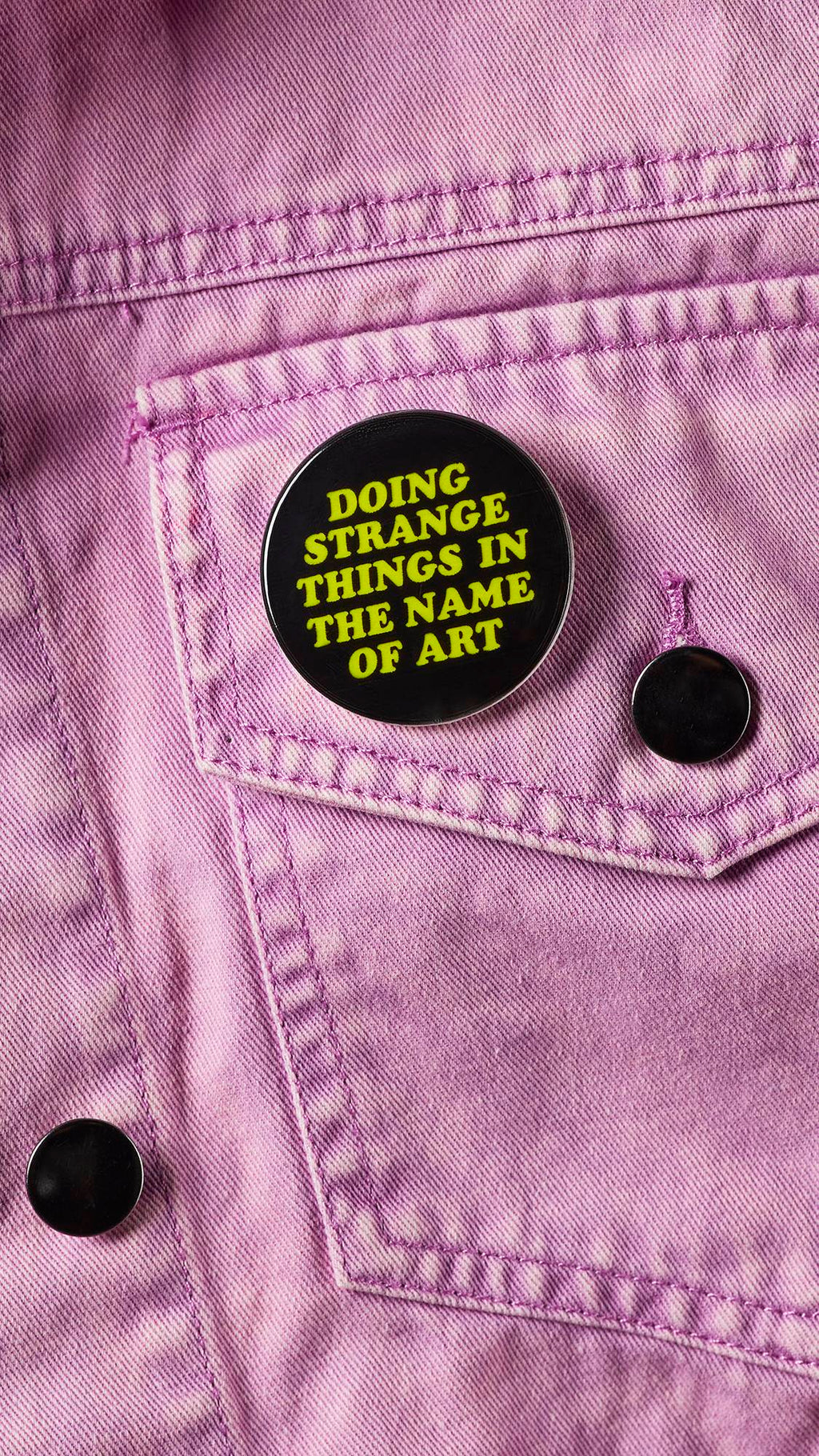 A close-up of the black badge with the green text "Doing Strange Things In The Name Of Art" pinned to a baby pink denim jacket pocket.