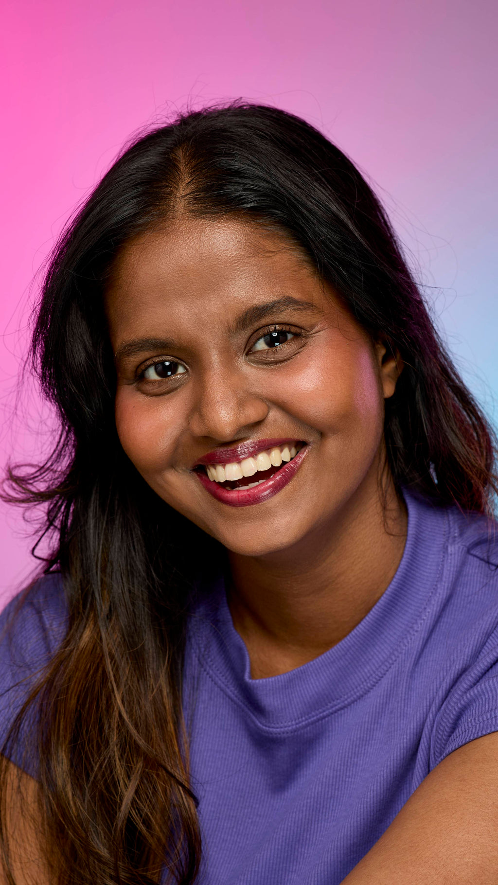 A smiling person with dark hair and skin, wearing a purple top, is shown from the chest up. They are wearing the Lush, deep red Dragon's Blood lip cream on a pink and purple gradient background.