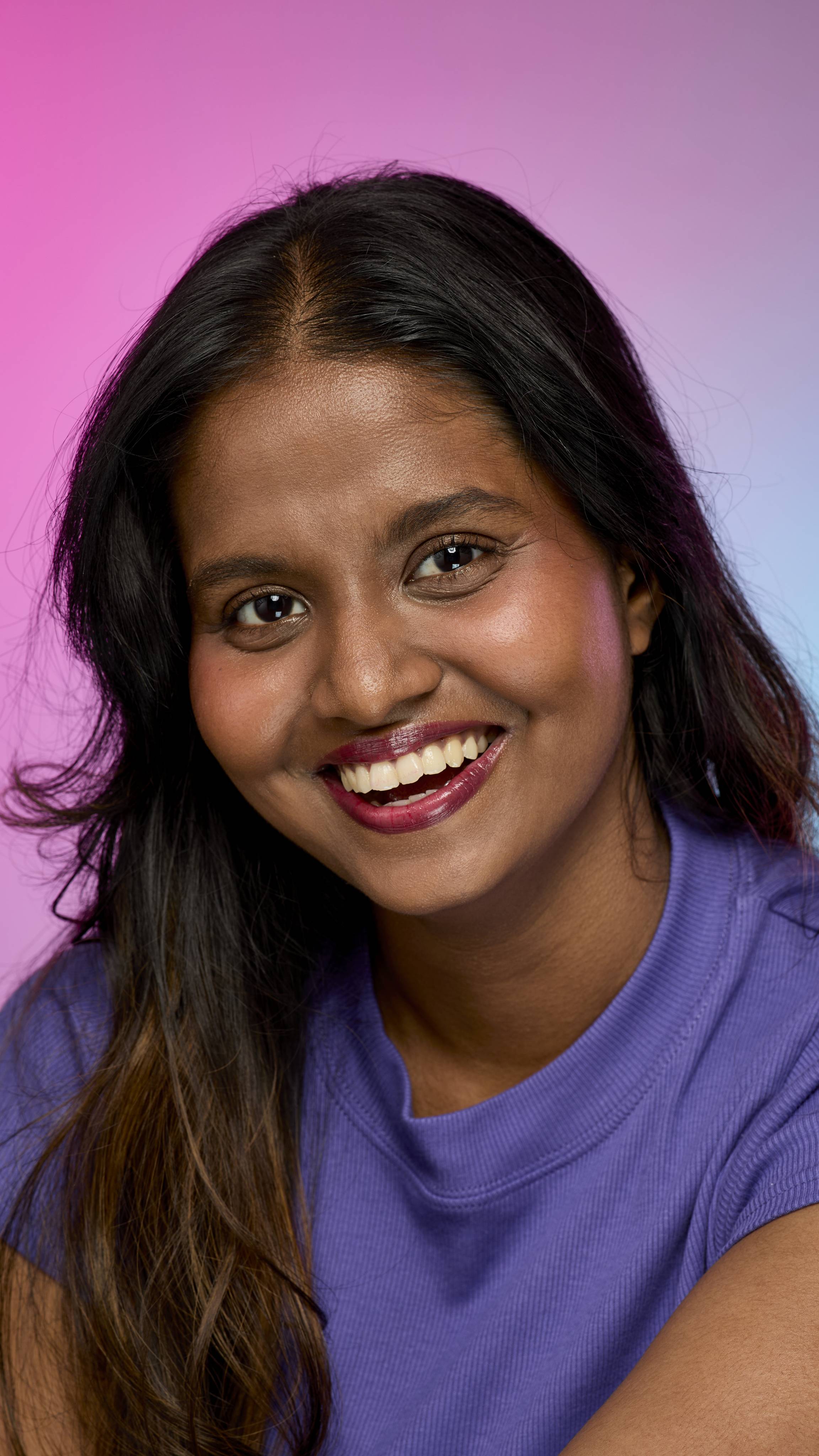 A smiling person with dark hair and skin, wearing a purple top, is shown from the chest up. They are wearing the Lush, deep red Dragon's Blood lip cream on a pink and purple gradient background.