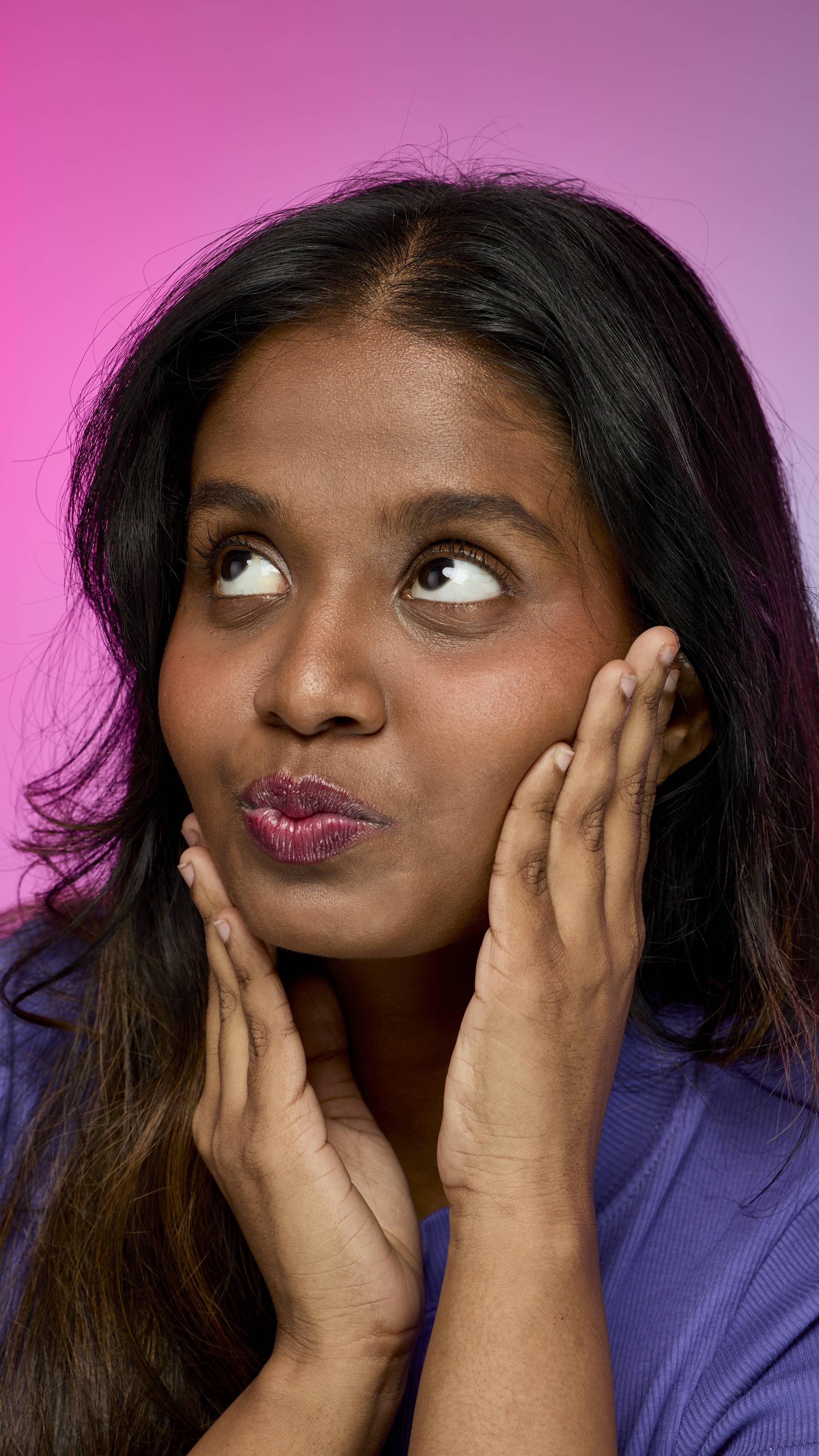 A smiling person with dark hair and skin, wearing a purple top, is shown from the chest up. They are wearing the Lush, deep red Dragon's Blood lip cream while making a pouty face on a pink and purple gradient background.