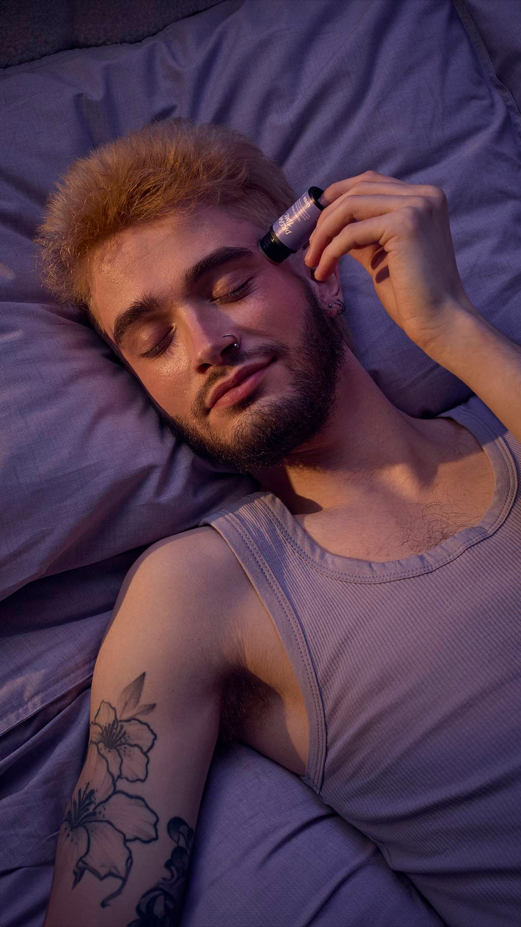 The image shows a close-up of the model's head as they are lying in bed in lavender-purple sheets. They are gently applying the balm directly to their temple as their eyes are closed in relaxation.