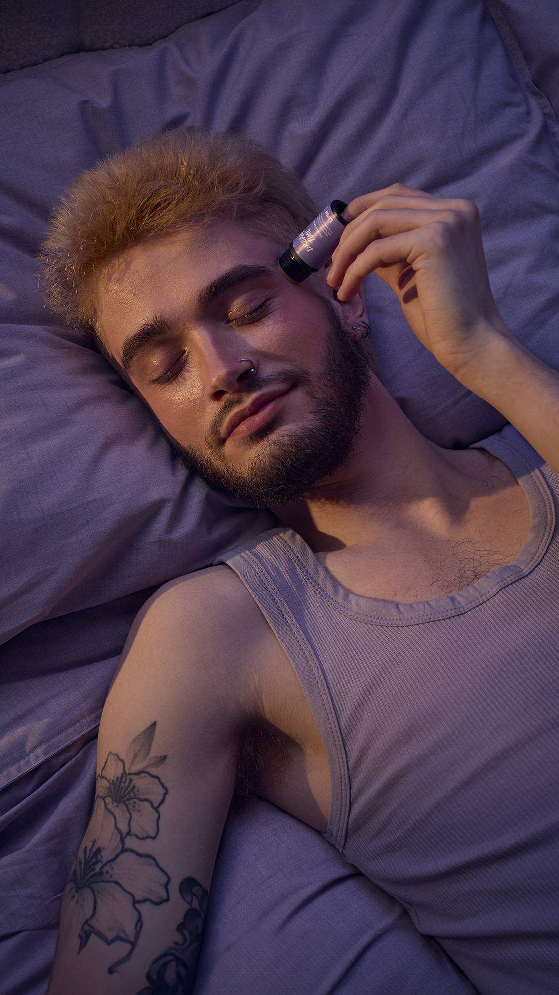 The image shows a close-up of the model's head as they are lying in bed in lavender-purple sheets. They are gently applying the balm directly to their temple as their eyes are closed in relaxation.