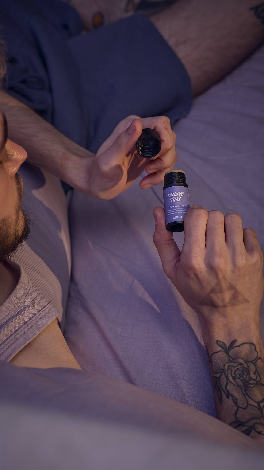 The image shows a close-up of the model as they are lying in bed in lavender-purple sheets. They have just taken the back lid off of the Temple Balm and are reading the front label.