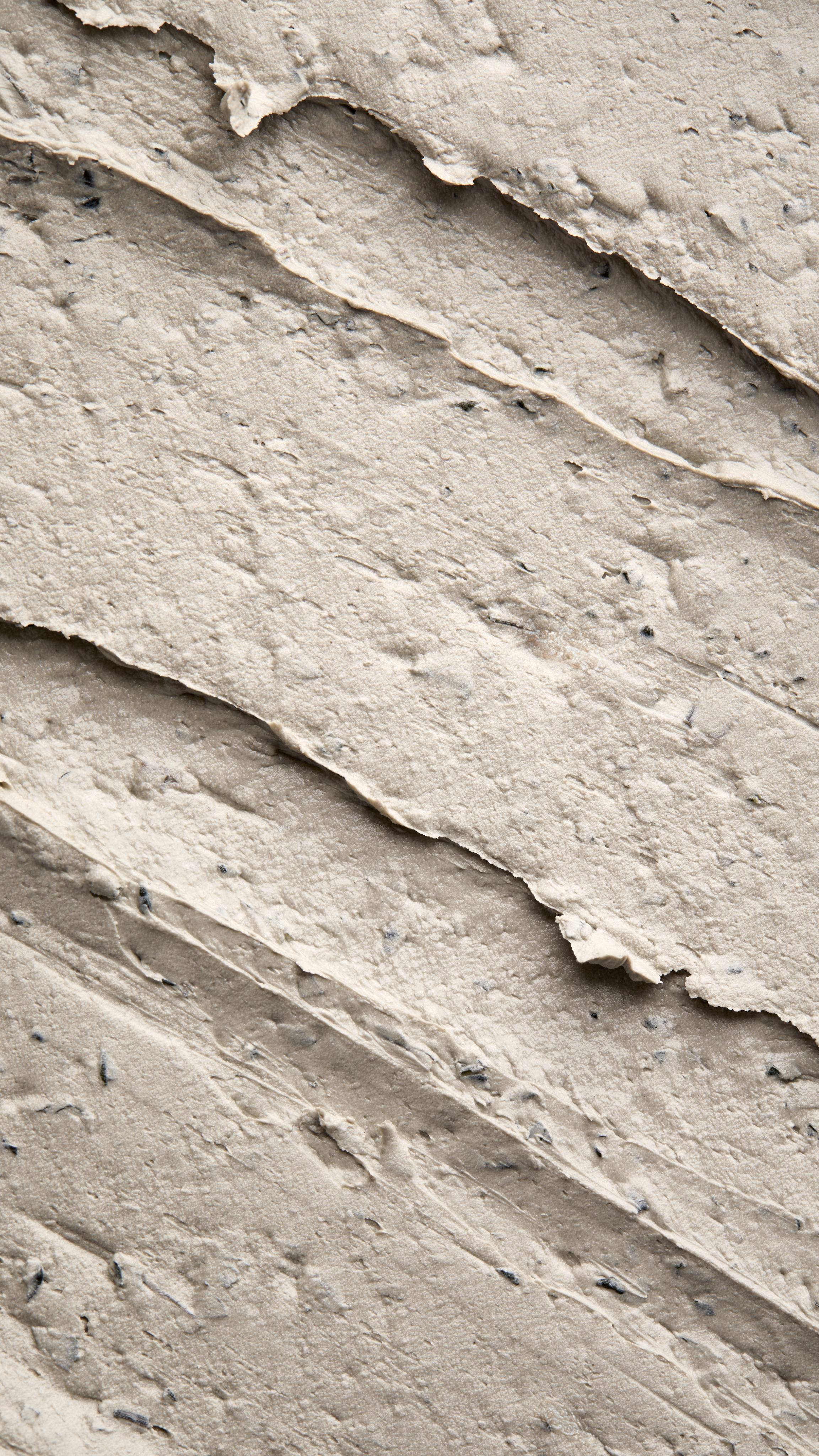 A close-up, textured shot of a thick, "Earthy, Woody Karma" foot mask spread in diagonal ridges. The product has a dense, matte consistency with visible dark exfoliating flecks and small botanical particles throughout.