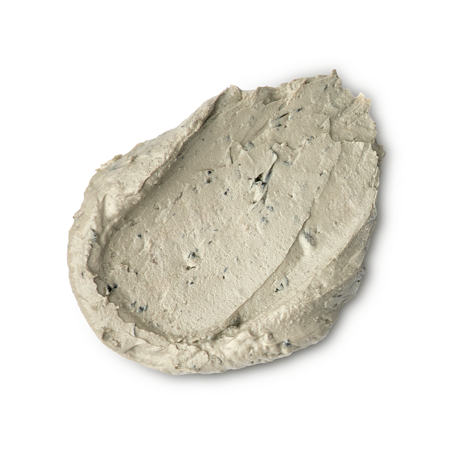 Earthy, Woody, Karma. A thick, textured dollop of pale beige foot mask. The product has a dense, matte consistency with visible dark exfoliating flecks and small lumps, showing a swirled and slightly peaked surface.