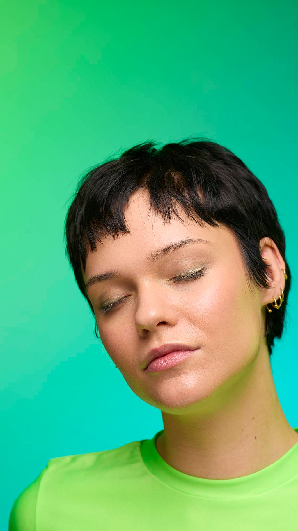 The image shows a close-up of the model's face on a pale blue-to-green gradient background. The model's make-up is done with the focal point being a subtle, gunmetal-brown, shimmering eyeshadow made by the Eclipse Glitter Crumble.