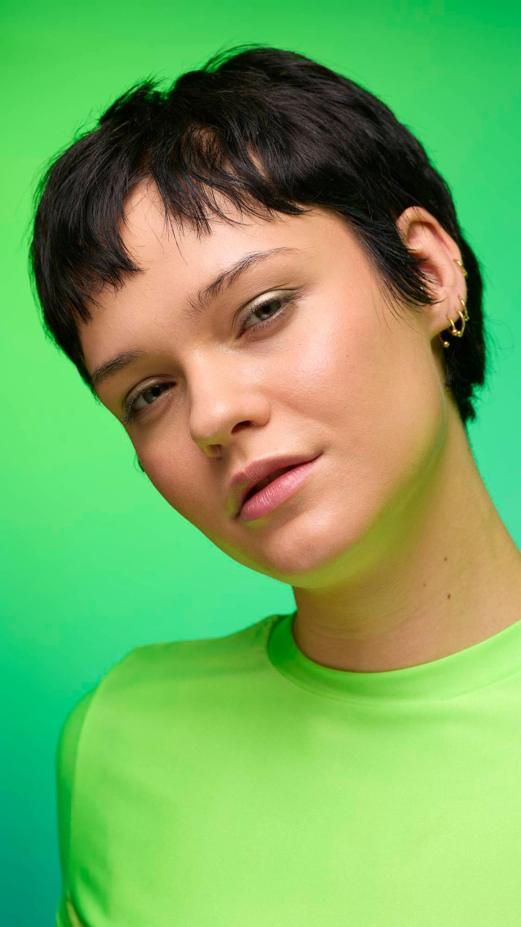The image shows a close-up of the model's face on a pale blue-to-green gradient background. The model's make-up is done with the focal point being a subtle, gunmetal-brown, shimmering eyeshadow made by the Eclipse Glitter Crumble.