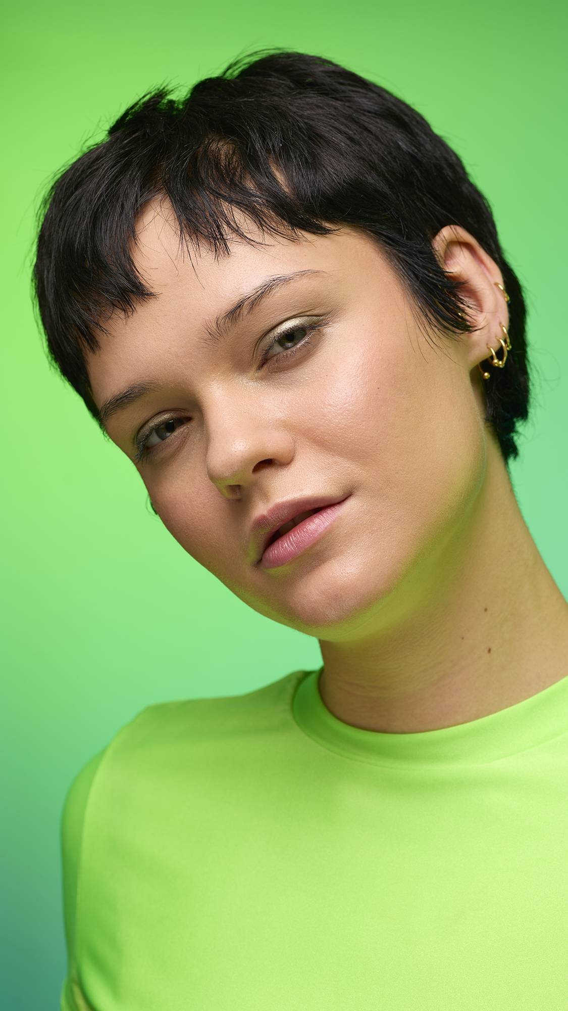 The image shows a close-up of the model's face on a pale blue-to-green gradient background. The model's make-up is done with the focal point being a subtle, gunmetal-brown, shimmering eyeshadow made by the Eclipse Glitter Crumble.