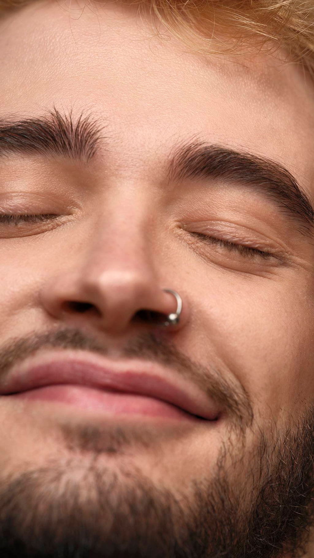 The model is on an earth-toned, warm background. They have their eyes closed as they are smiling at the camera with glowing, freshly moisturised skin and bright under-eyes.