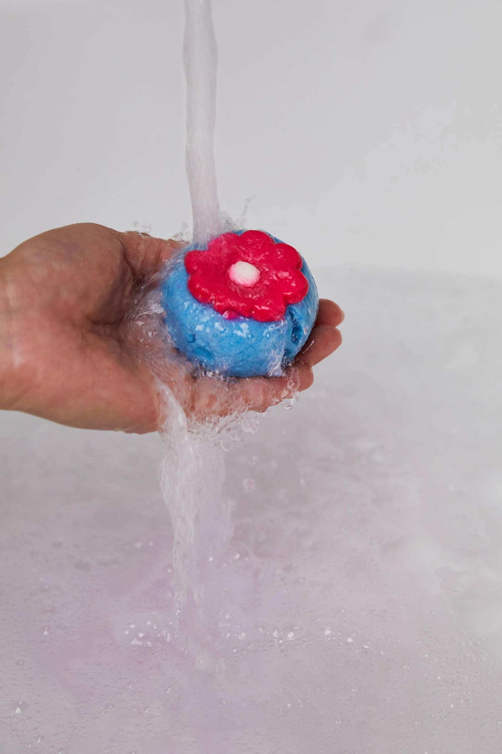 A close-up image of the model's hand reaching in from the left as they hold the blue with a pink flower, pressed sphere-shaped Pop In The Bath bubble bar under running bath water. The product creates a layer of thick bubbles in the bath below.