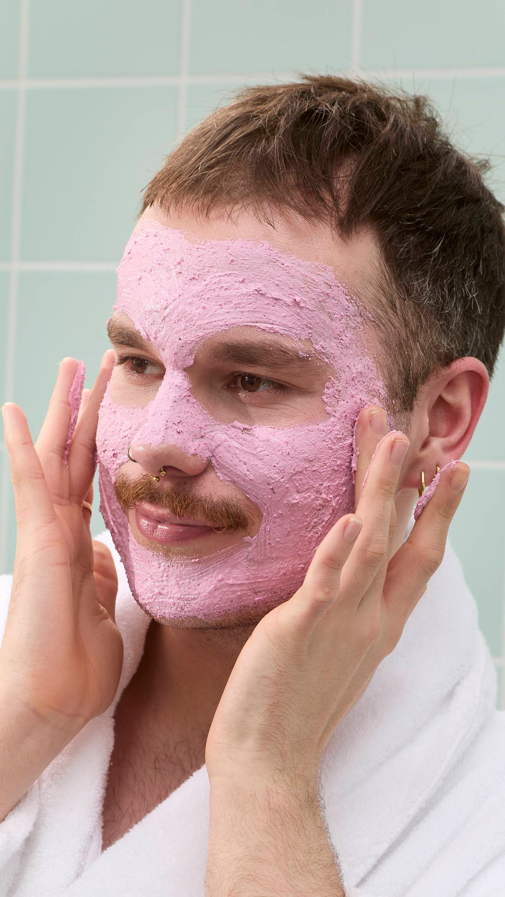 A model with a moustache and nose piercings smiles while looking to the side. Most of their face is covered in a thick, pink "Family Face Time" face mask. They wear a white dressing gown against a background of pale green bathroom tiles.