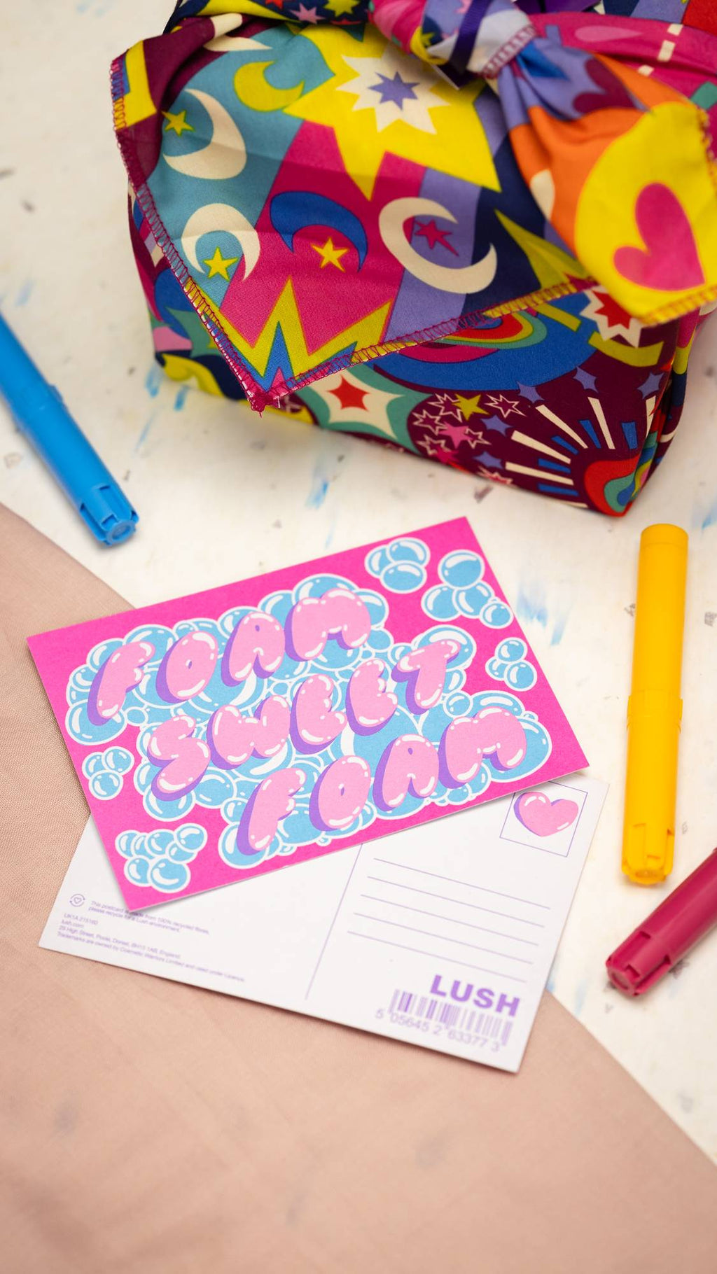 Image shows the Foam Sweet Foam postcard laid on the table next to a pen and a LUSH knot-wrapped gift.