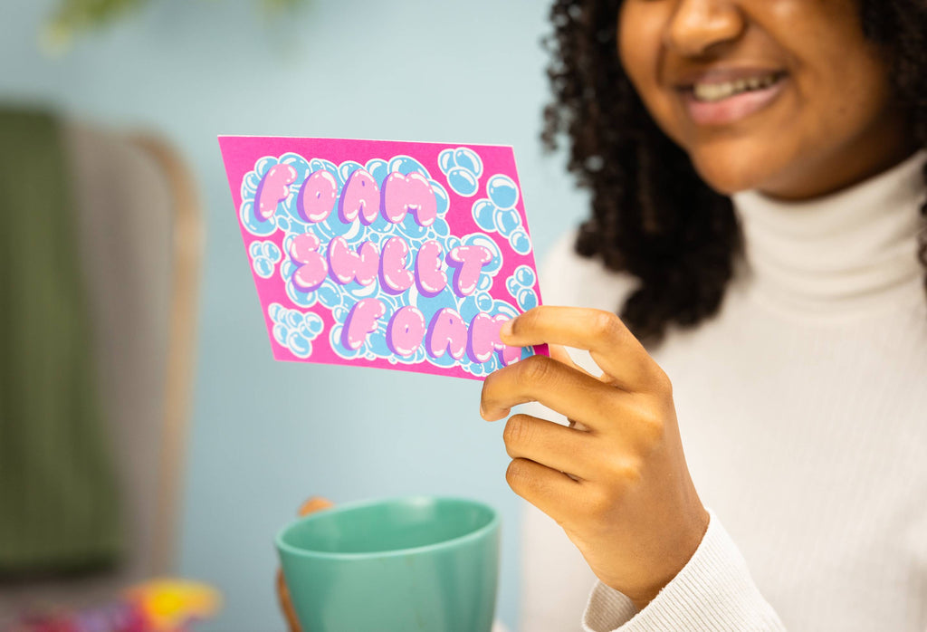 Image shows the model holding the Foam Sweet Foam postcard as they read it with a cup of tea.