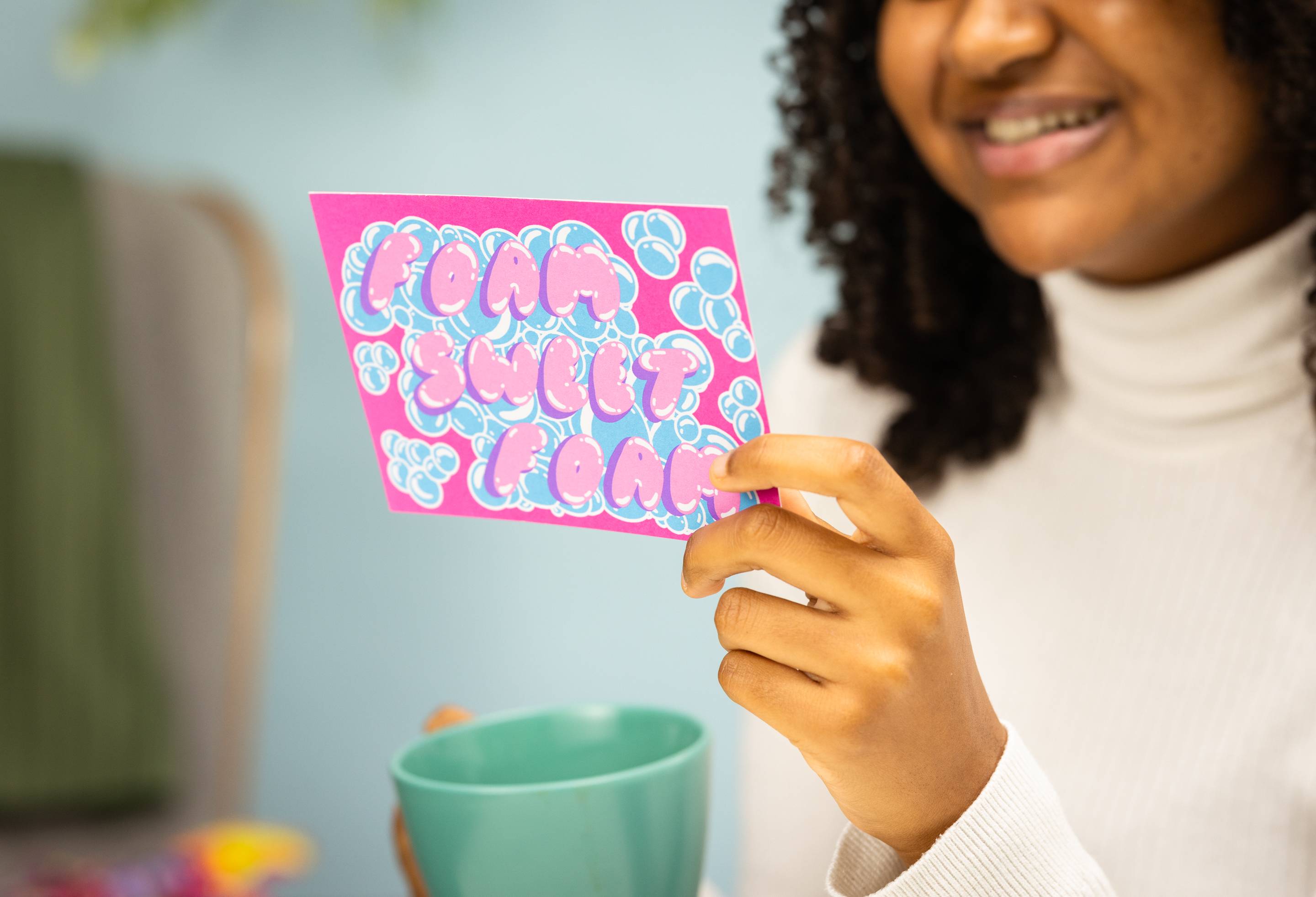 Image shows the model holding the Foam Sweet Foam postcard as they read it with a cup of tea.