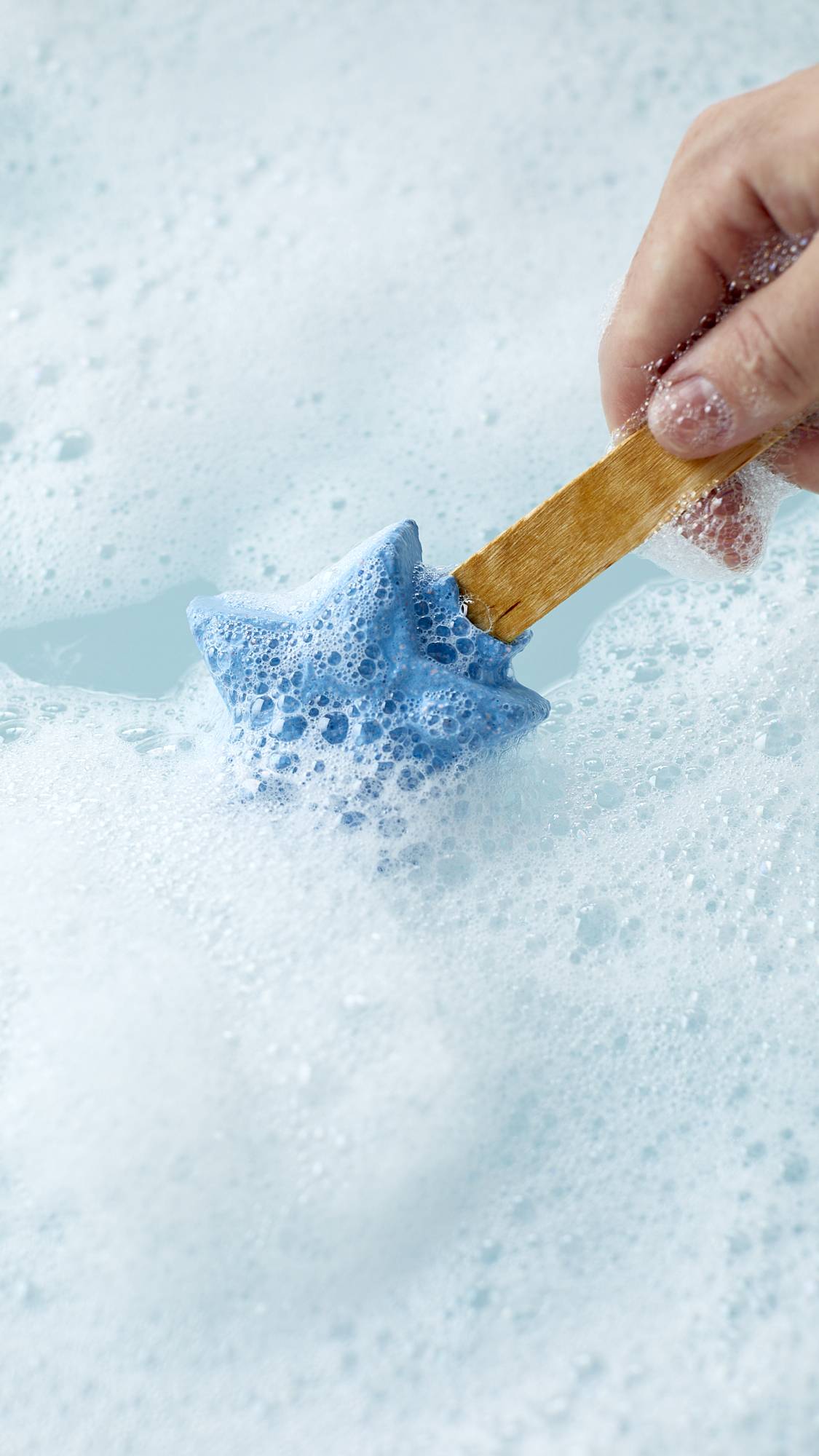 The image shows a close-up shot of the Frozen reusable bubble bar stick being swirled around cool, icy-blue bath water surrounded by a thick blanket of foamy bubbles made by the product.