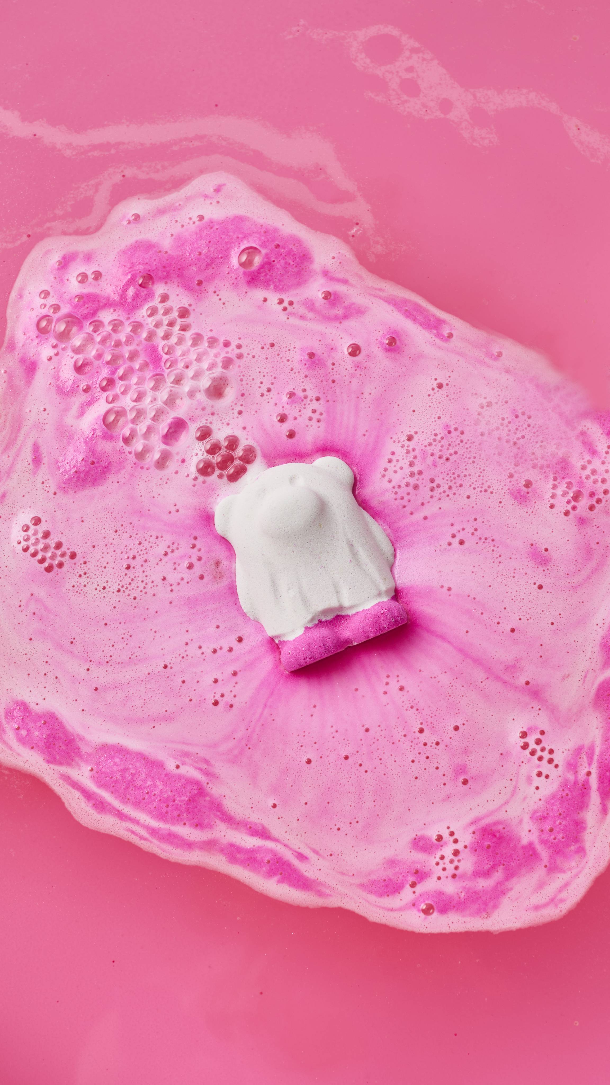 A white ghost bear bath bomb fizzes and dissolves in bright pink bathwater, floating in a pool of vibrant pink and white foam and bubbles.