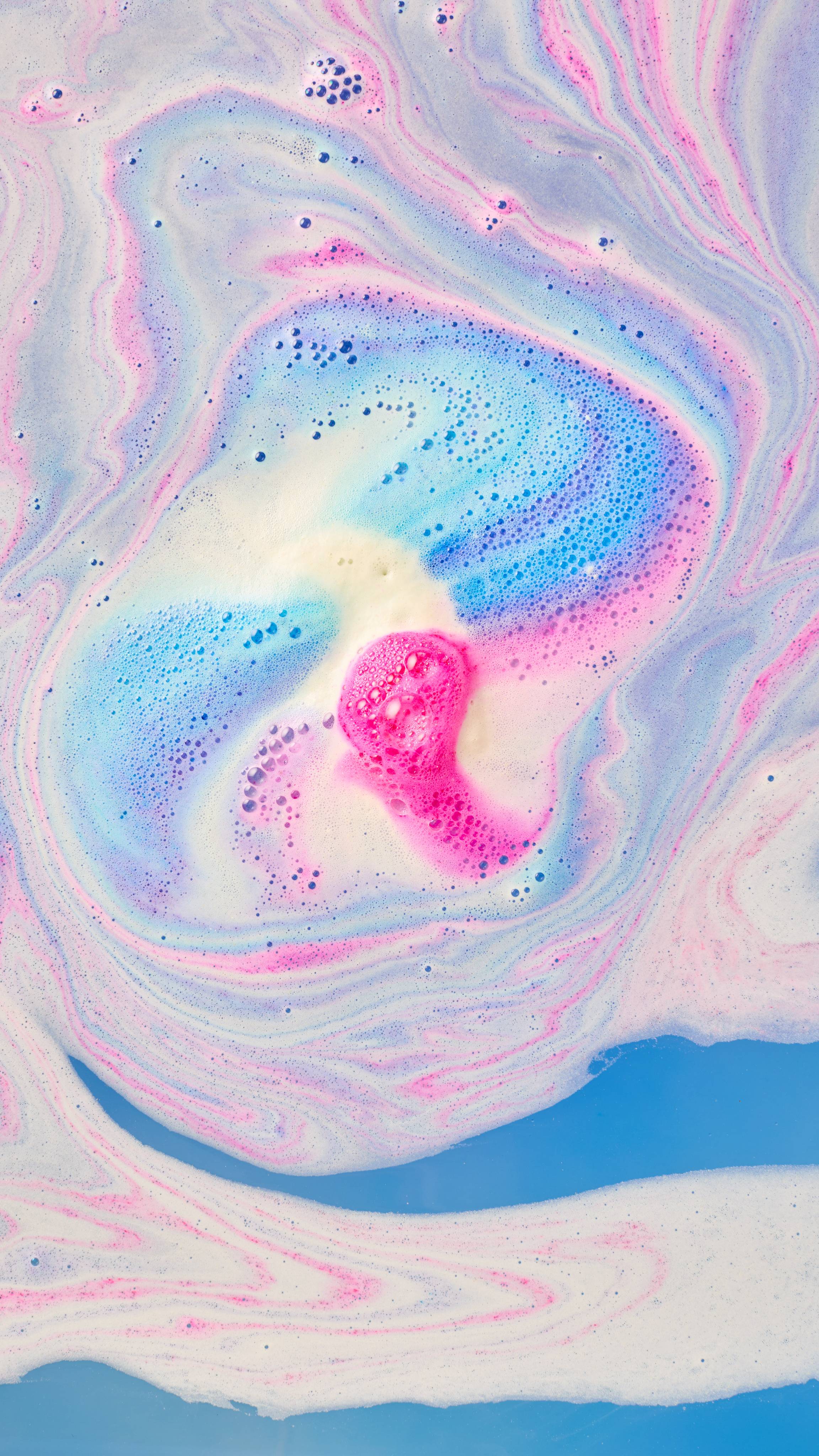 A vibrant swirl of hot pink and bright blue foam mixes in the centre of a bath, surrounded by marbled pastel ripples of white, pink, and violet froth. Clear blue water is visible at the bottom.