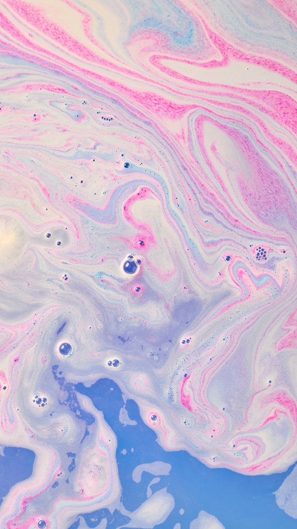 An abstract aerial shot of swirling bath bomb foam in water, forming marbled patterns of bright pink, violet, and pale cream. Deep blue bath water with scattered soapy bubbles is visible at the bottom.