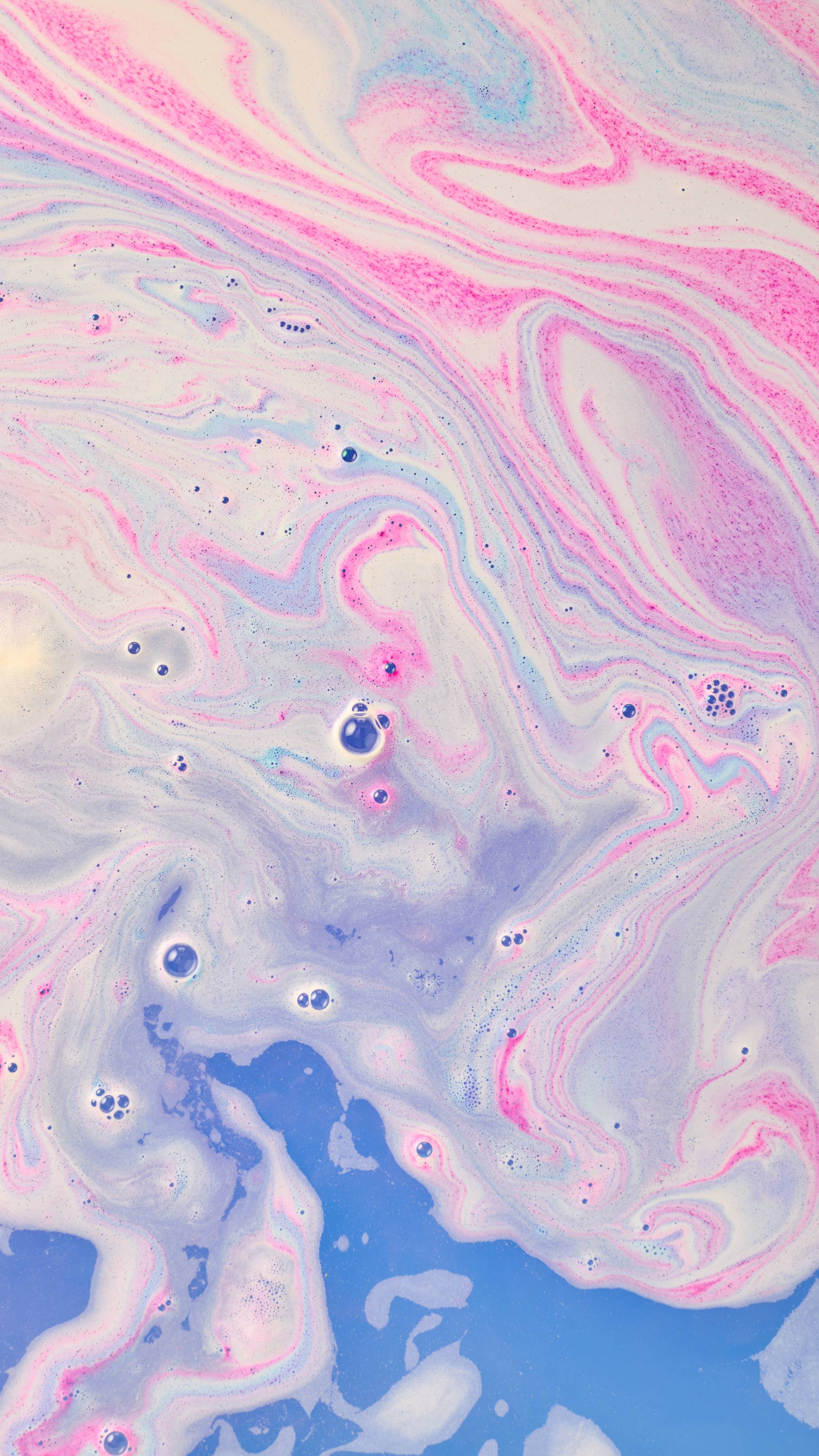 An abstract aerial shot of swirling bath bomb foam in water, forming marbled patterns of bright pink, violet, and pale cream. Deep blue bath water with scattered soapy bubbles is visible at the bottom.