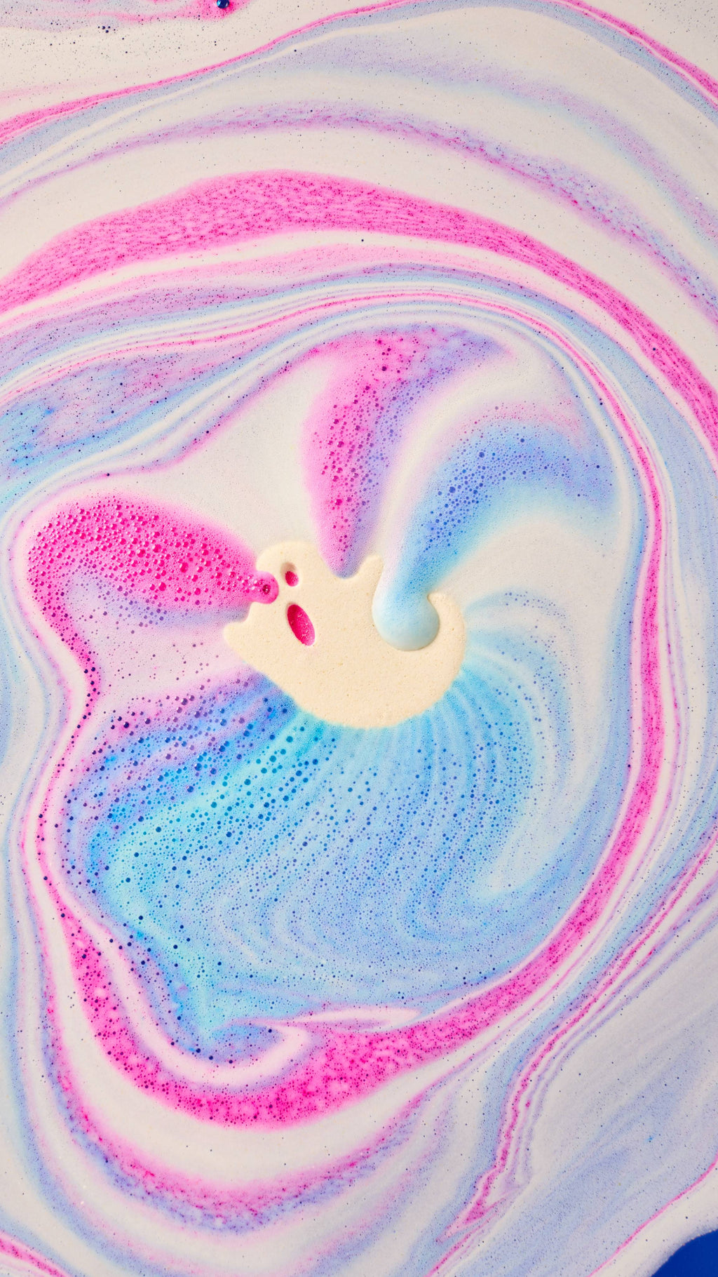 A cream-coloured, ghost-shaped bath bomb dissolves at the centre of a bath, surrounded by swirling patterns of bright pink, blue, and violet foam that ripple across the water's surface.