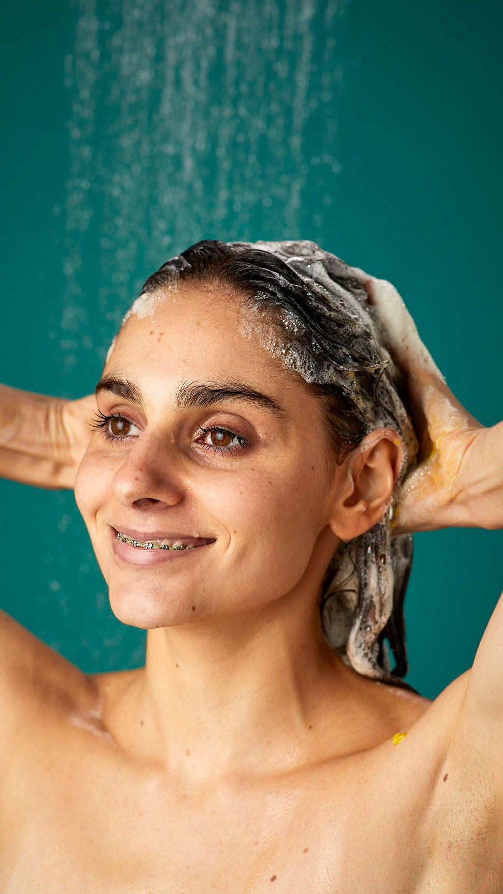 The model is standing under running shower water on a teal background. Their dark, long hair is wet and lathered in suds as they lather the Ginger shampoo into their roots.