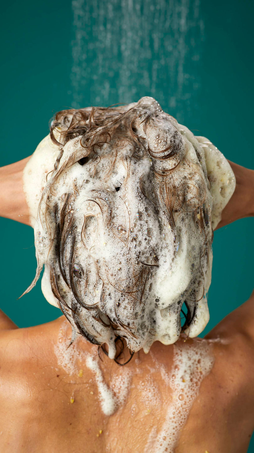 The image shows the rear view of the model's dark, long hair which is wet and lathered in suds as they lather the Ginger shampoo into their roots. There is a teal background with running shower water.