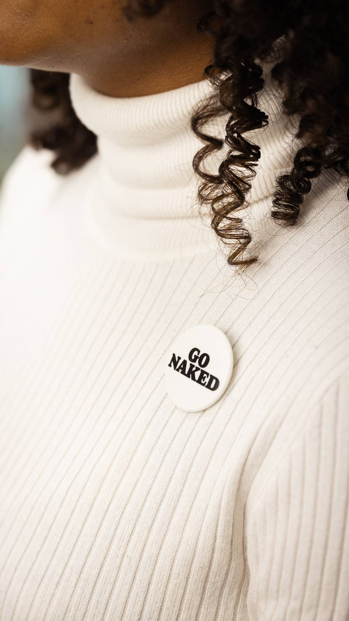 A close-up of the model wearing a white turtle neck with dark, curly hair. They are wearing the "Go Naked" badge.