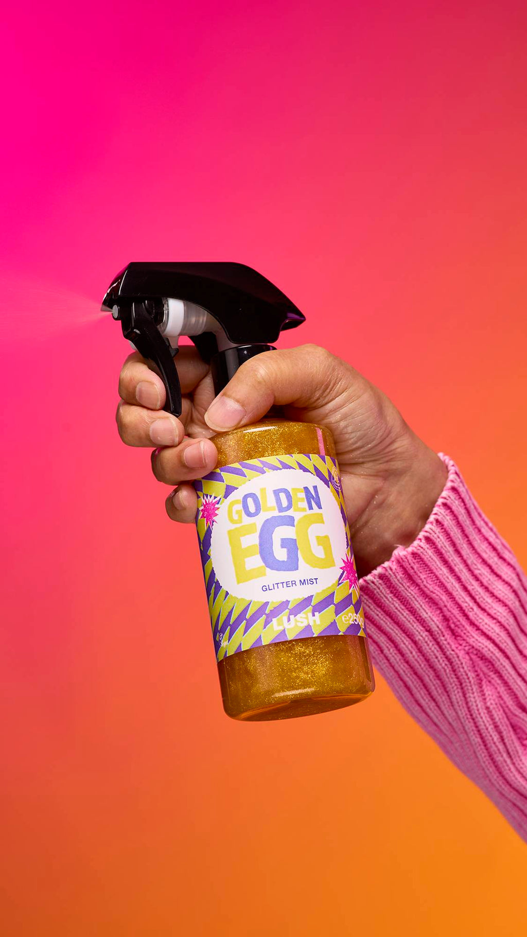 An arm is reaching from the right-hand side. The hand holds a transparent, cylindrical plastic spray bottle with golden, shimmering liquid and a black top. A colourful label reads "Golden Egg Glitter Mist" on a pink-to-orange gradient background.