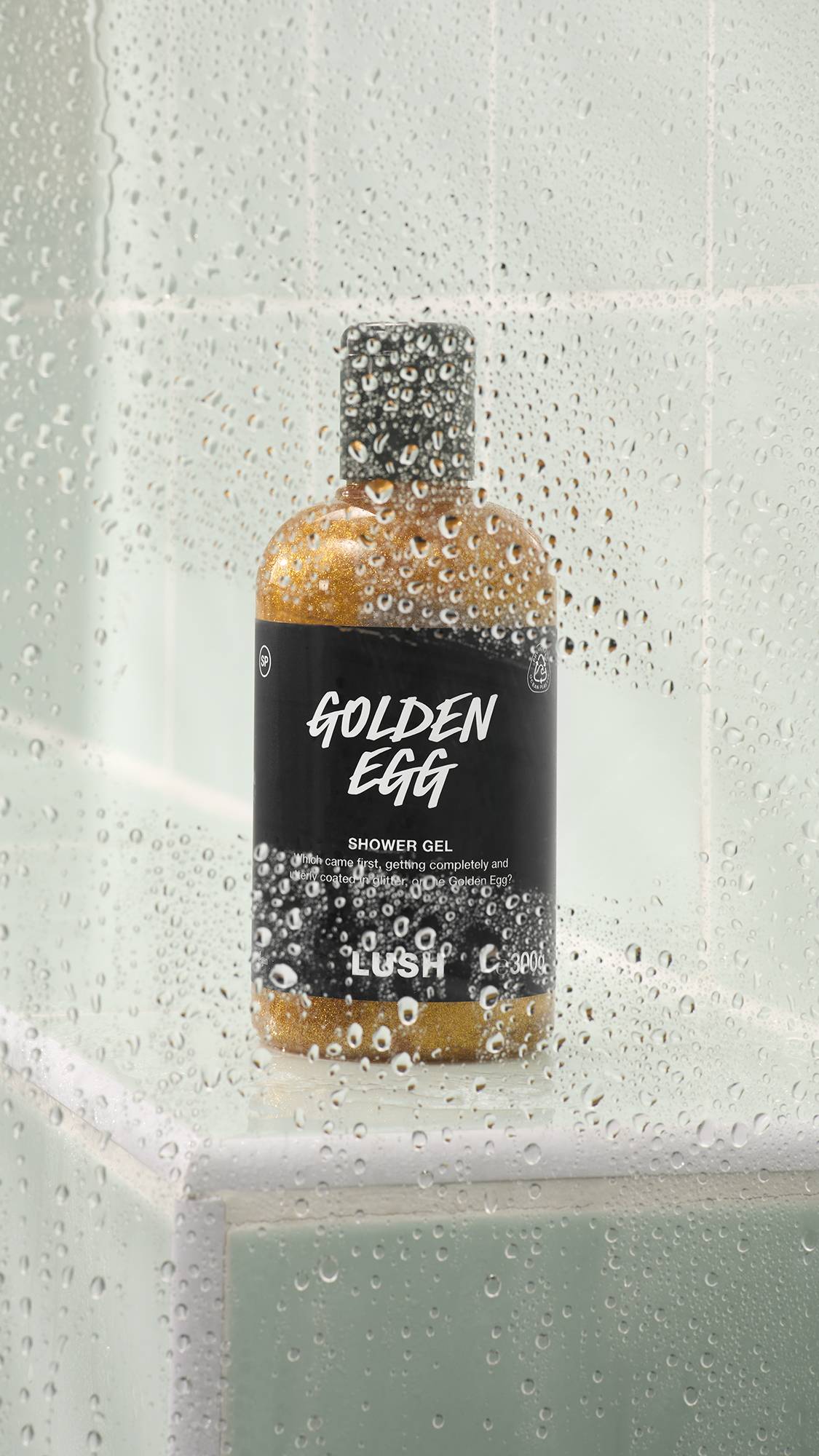 A bottle of Lush Golden Egg shower gel filled with shimmering gold glitter liquid sits on a tiled shower ledge. The view is partially obscured by a glass shower door covered in clear water droplets.