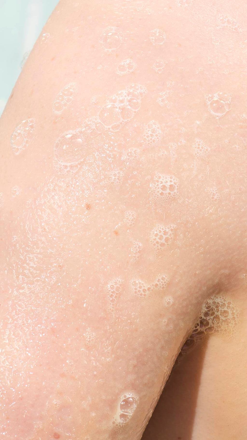 A close-up of a person's skin in the shower, covered in a thin layer of shimmering gold-tinted soap suds. Small, delicate white bubbles are scattered across the damp skin, creating a glistening, hydrated texture.